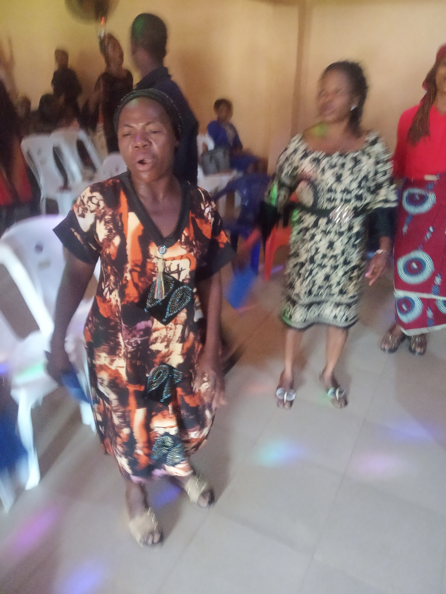A medium-shot photograph shows a group of women dancing in what appears to be a room or hall, likely in Nkwelle Ezunaka, Nigeria. The lighting is somewhat dim with a few scattered colorful lights.
The woman in the foreground is wearing a patterned dress with short sleeves and embellishments and is singing. Her mouth is open. Next to her to the right is another woman, also in a patterned dress, belted at the waist. To the right of her is another woman in a red top and patterned skirt. All of the women are wearing sandals.
There are white plastic chairs visible on the left, and blurred figures of other people in the background. The background is out of focus.