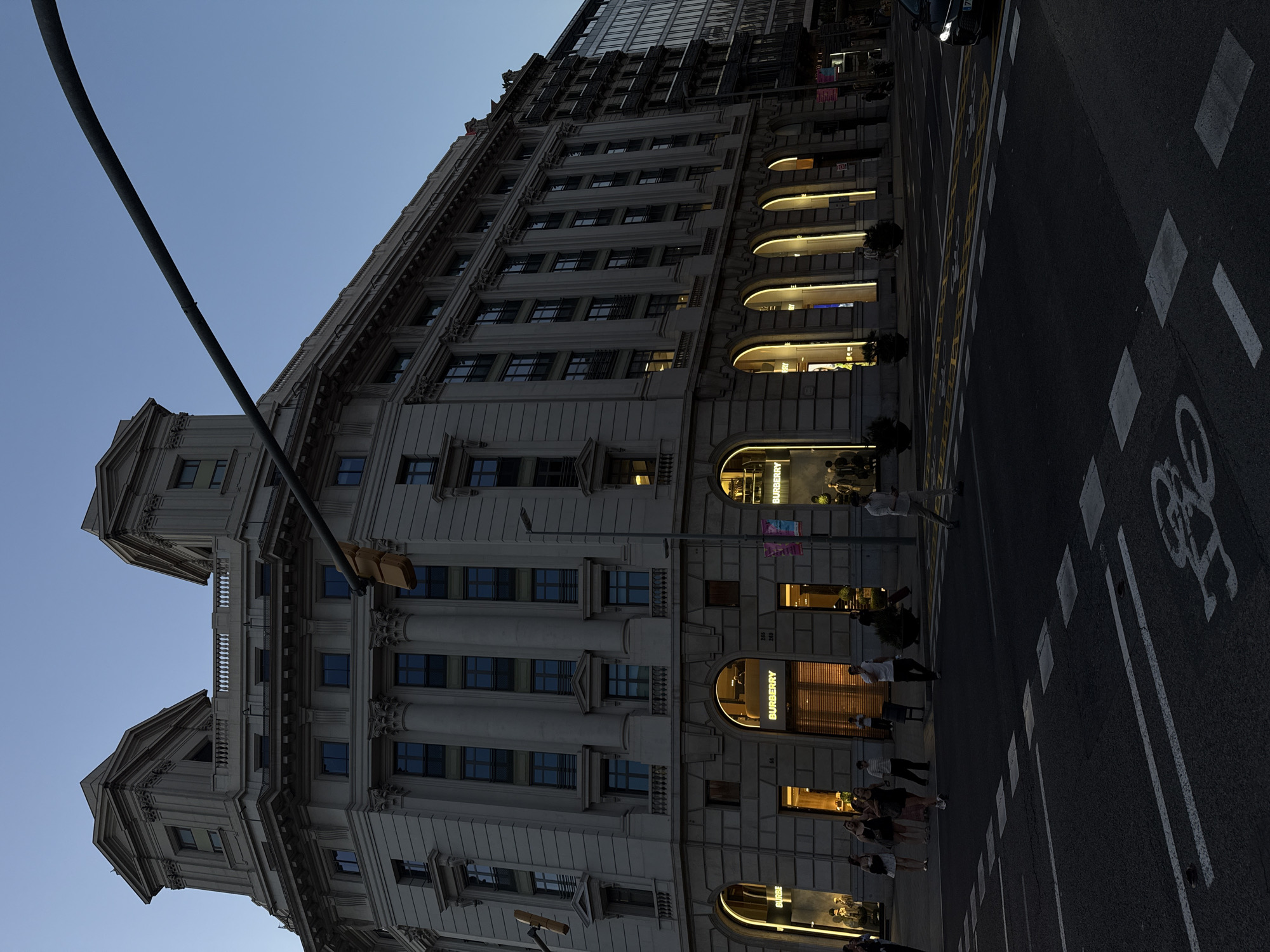 This media file captures an evening scene in Barcelona, Spain, focusing on a grand, ornate stone building that stretches along an urban street. The image is taken at dusk or early evening, characterized by a deep blue sky and artificial illumination from streetlights and shopfronts.
The dominant subject is a multi-story building featuring classical architecture with columns, decorative cornices, and numerous windows. The lower levels showcase several arched shopfronts, brightly lit from within. Prominently displayed are multiple "BURBERRY" signs, indicating the presence of a luxury fashion retailer. Near one of these signs, the numbers "255" and "259" are visible. Above the stone structure, a modern, darker glass facade extends upwards, suggesting a newer addition or a different section of the building.
In the foreground, a dark asphalt road is visible, marked with white dashed lines, yellow lines, and a white painted bicycle symbol, indicating a dedicated lane for cyclists. Several pedestrians are scattered along the sidewalk in front of the shops, some appearing to be browsing or walking. Their forms are somewhat blurred, suggesting movement or low light conditions. A dark, curved pole or streetlight extends diagonally across the top left portion of the sky. The headlights of a car are visible in the upper right corner, partially obscured. The scene suggests a bustling commercial area coming alive as evening falls.