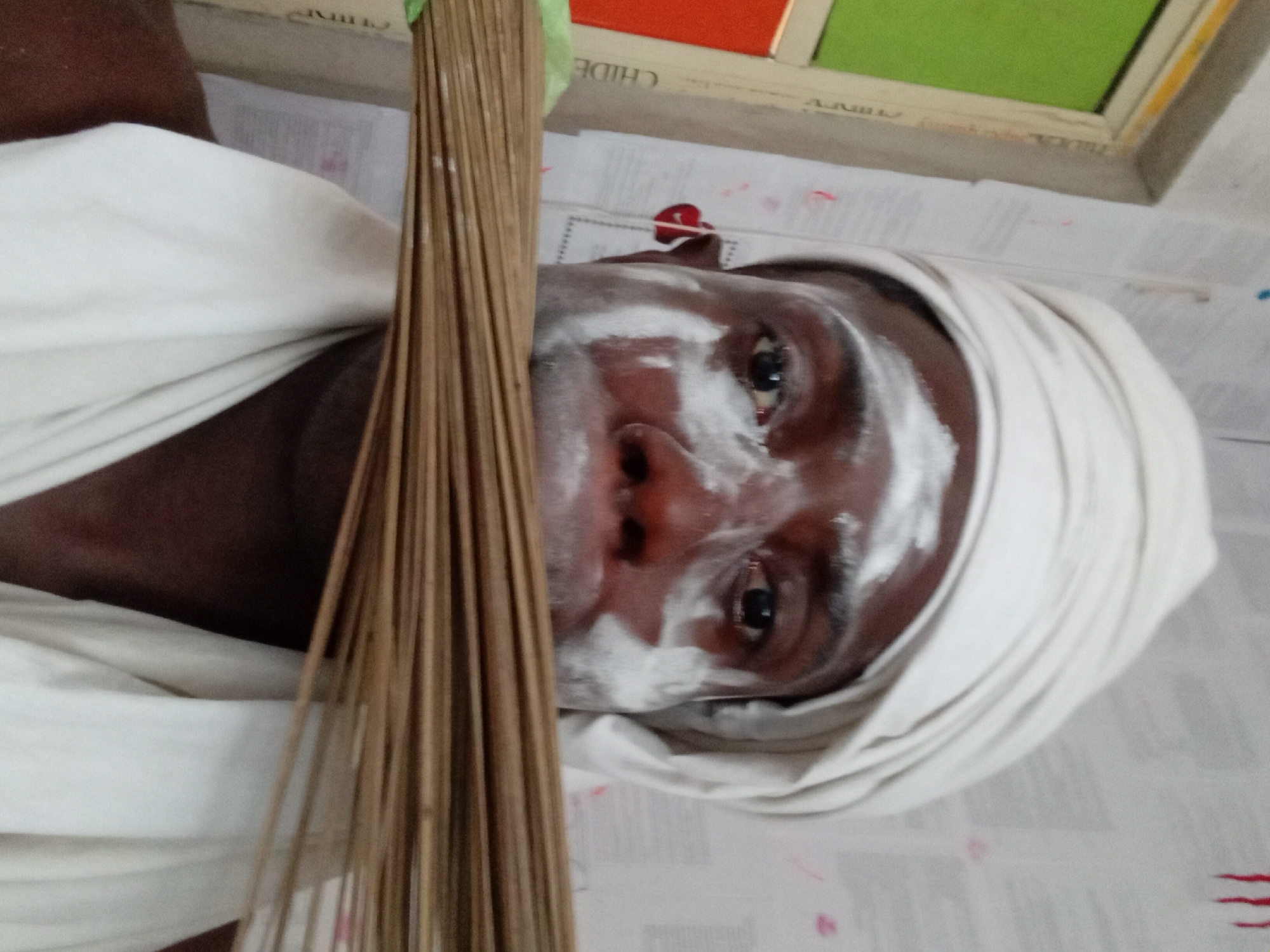 A close-up, tilted shot of a person with dark skin partially covered with white cream or paint on their face. The person is also wearing a white cloth wrapped around their head. A bundle of thin, brown sticks is positioned in the foreground, partially obscuring the person's face. The background appears to be a surface covered with paper or print, possibly a newspaper. A window frame with sections of green and orange is visible in the upper part of the image. The location is Akwete, Nigeria.