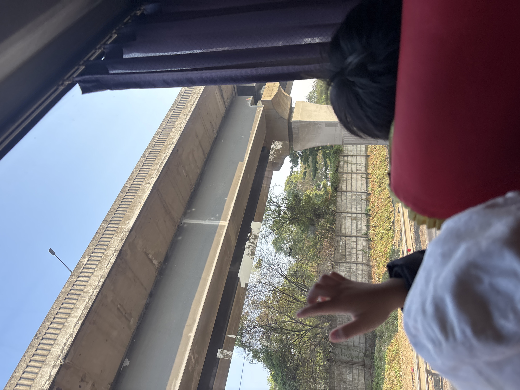 The image shows a view from inside a vehicle, likely a bus or train, looking out a window. A person's hand is visible in the foreground, raised with index finger extended. Beyond the hand, a concrete overpass stretches across the frame, supported by large concrete pillars. Trees and a concrete wall line the area beneath the overpass. In the distance, train tracks are visible. The sky is clear and blue. A dark purple curtain hangs on the right side of the window. The scene suggests travel through an urban or suburban environment, possibly Karawang, Indonesia, given the provided context.
