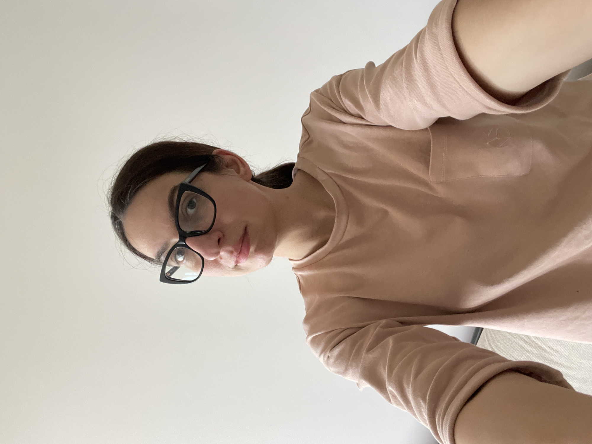 A person is seen from a high angle looking down at the viewer, wearing black-framed glasses and a light pink long-sleeved shirt. The background is a plain white or light-colored surface. No elements indicate a location in Shishmantsi, Bulgaria.