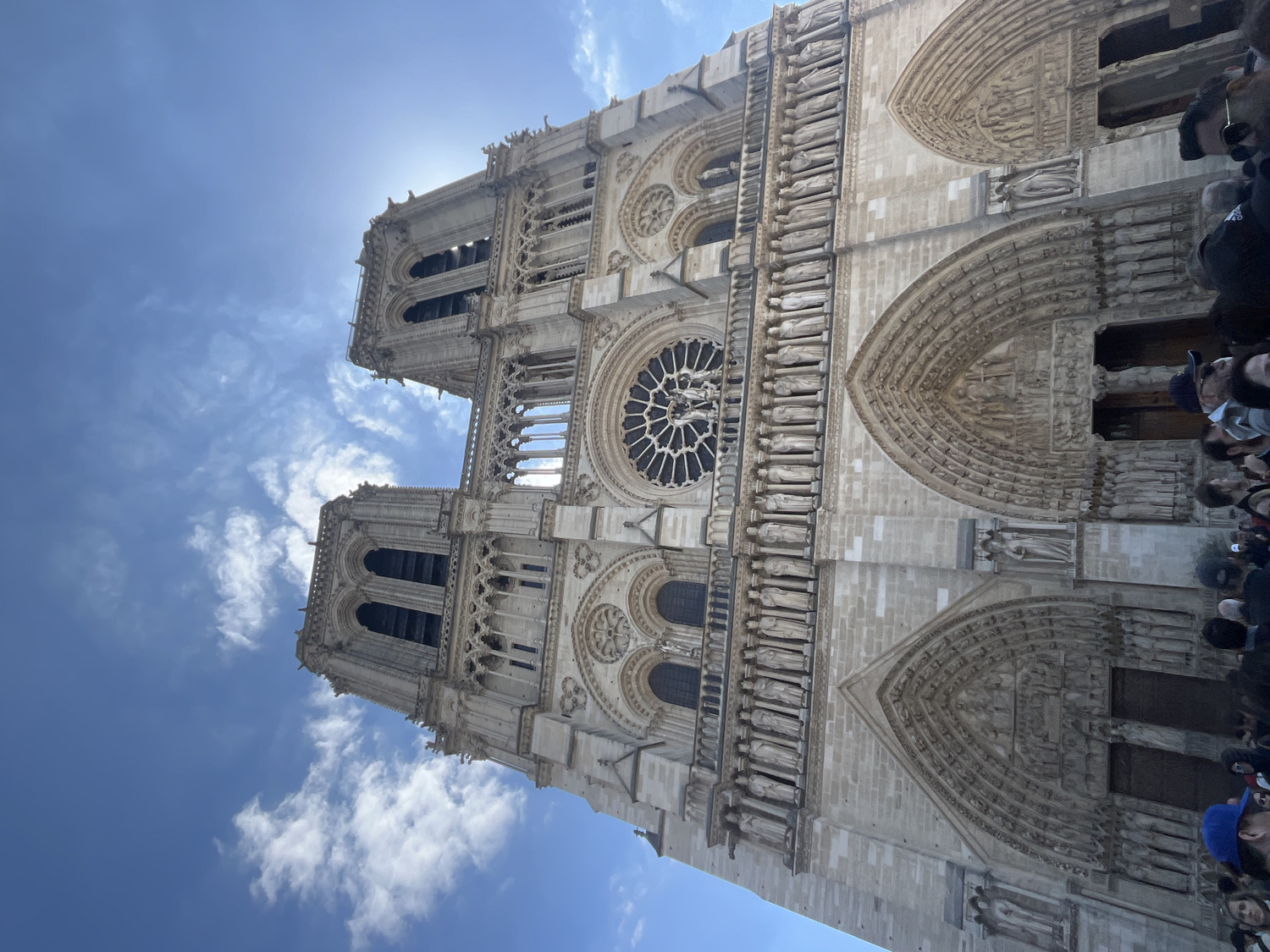The image captures a striking, low-angle view of the facade of Notre Dame Cathedral in Paris, France, rotated 90 degrees clockwise. The monumental Gothic architecture dominates the frame, showcasing its intricate details under a bright, sunny sky.
The scene is set outdoors during daylight hours, likely mid-day or early afternoon, given the high position of the sun, which peeks from behind the upper right tower, casting a slight glare. The sky is a vibrant blue with scattered white, wispy clouds, indicating clear and pleasant weather.
The primary subject is the cathedral's ornate stone facade, featuring its two iconic square towers at the top. Below these, a large, detailed rose window with elaborate tracery is prominently centered. Rows of sculptures, including the Gallery of Kings, are visible across the mid-section. At the lower level, three large arched portals are adorned with intricate carvings and sculptural tympana, leading into the cathedral. The stone appears light-colored, reflecting the sunlight.
A crowd of people is visible in the lower right portion of the image, standing directly in front of the cathedral. They appear to be tourists or visitors, looking up at the grand structure. Their faces are mostly obscured, but their presence indicates an active sightseeing or visitation event. No specific activities beyond observation are discernible. No discernible text is visible on the building or among the crowd.