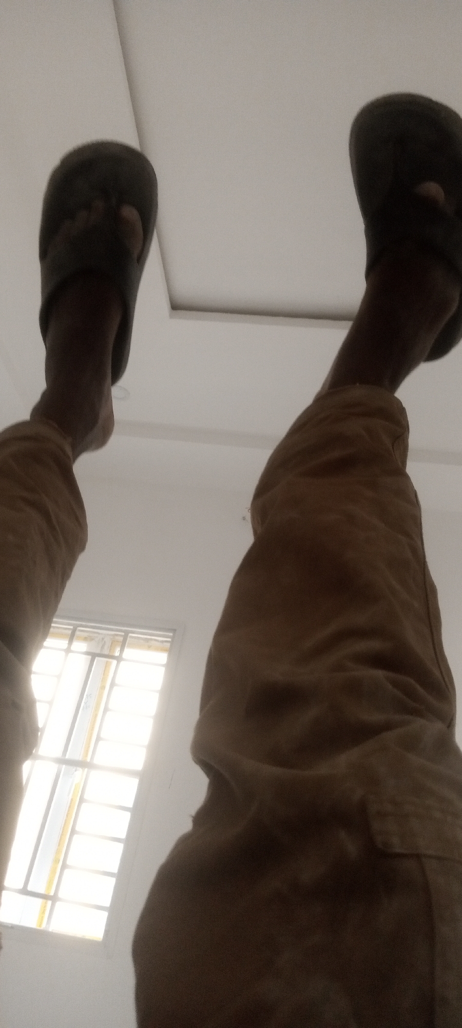 A low-angle shot depicts a person's legs and feet wearing brown trousers and dark slippers. The person is standing inside a building, possibly in Badiko, Nigeria, suggested by the context. A window with a barred frame is visible in the lower left, and the ceiling features recessed lighting panels. The perspective suggests the photo was taken from a low position, looking upwards.