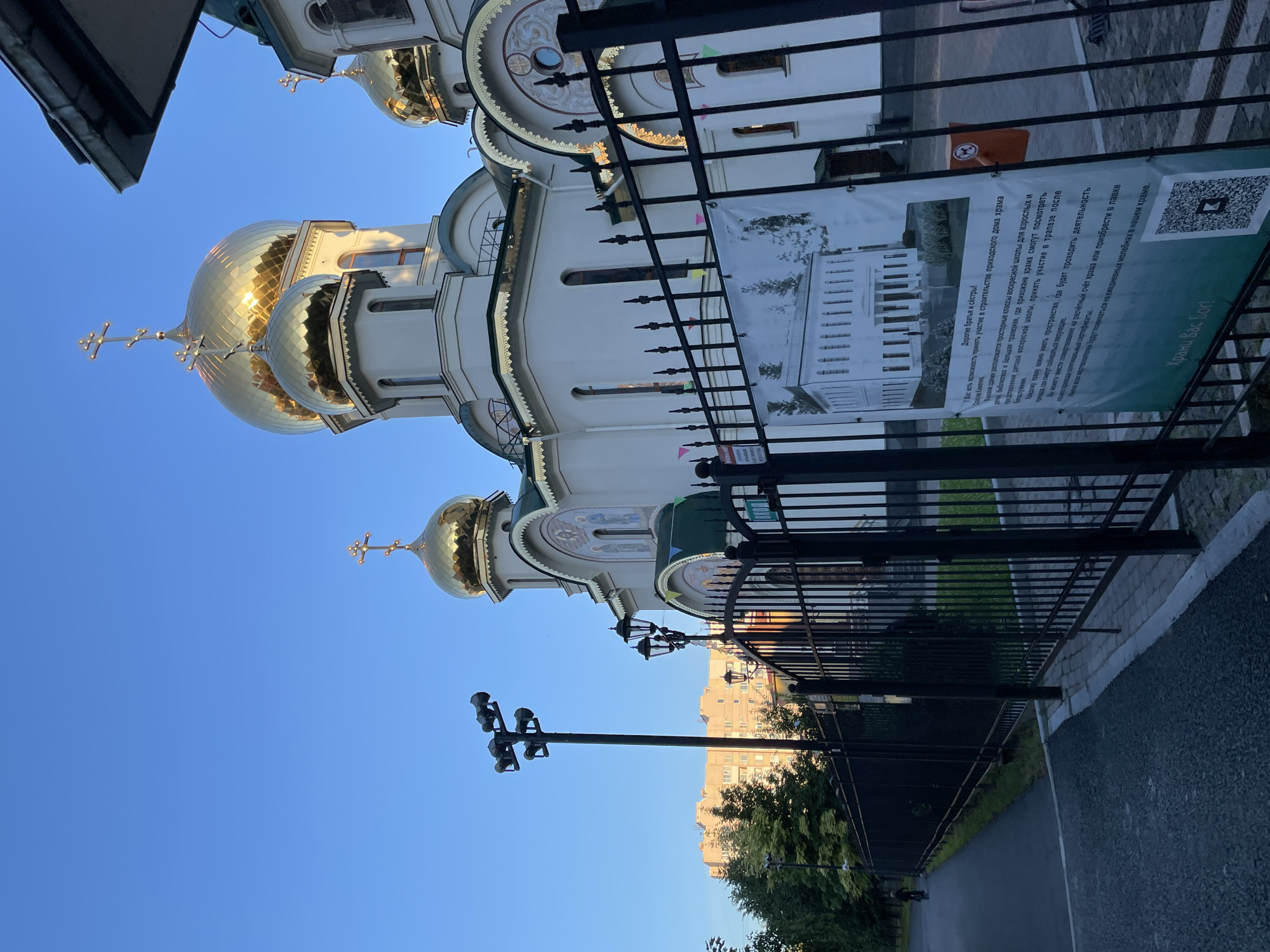 This image depicts the Church of the Resurrection of Christ, also known as the Smolensk Cemetery Church, located in Saint Petersburg, Russia. The exterior of the church, with its white walls and prominent golden domes topped with crosses, is visible under a clear blue sky. In the foreground, a black metal fence encloses the church grounds. A large banner is attached to the fence, displaying an architectural drawing of a building and text in Russian. Adjacent to the banner is an orange bin. To the left of the church, a paved walkway runs alongside the fence, with a lamppost and trees lining the path. In the distance, a multi-story apartment building can be seen through the trees. The scene is bathed in the warm light of what appears to be early morning or late afternoon.