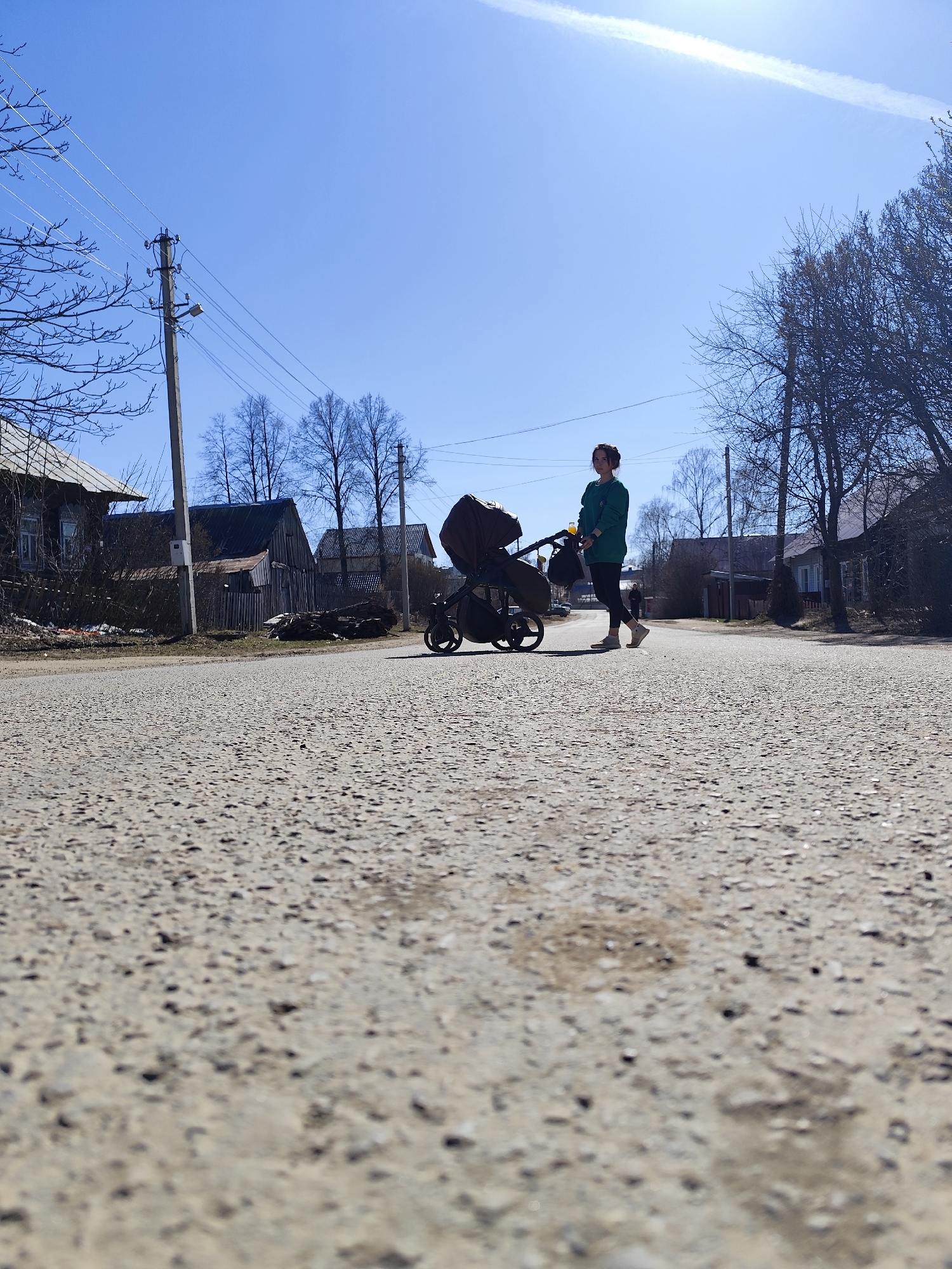 A low-angle outdoor photograph captures a woman pushing a baby stroller down a street in the village of Фоки, Russia. The woman is seen in profile on the right side of the frame, wearing a green long-sleeved top, dark leggings, and light-colored shoes. Her dark hair is pulled back. She pushes a dark, modern baby stroller with a bag hanging from its handle, containing a visible yellow bottle.
The street, occupying the foreground, is a textured, unpaved or gravel road. In the background, on both sides of the street, are several traditional wooden houses and structures, some appearing rustic. Bare-branched trees, indicative of early spring or late autumn, are scattered throughout the scene. A tall utility pole with numerous wires and a street light stands prominently on the left side of the road.
The sky is a clear, bright blue, suggesting a sunny day. A distinct white contrail stretches diagonally across the upper sky. The strong daylight implies it is daytime, likely morning or early afternoon. The scene portrays a calm, everyday activity in a rural or semi-rural setting. No text is visible in the image.