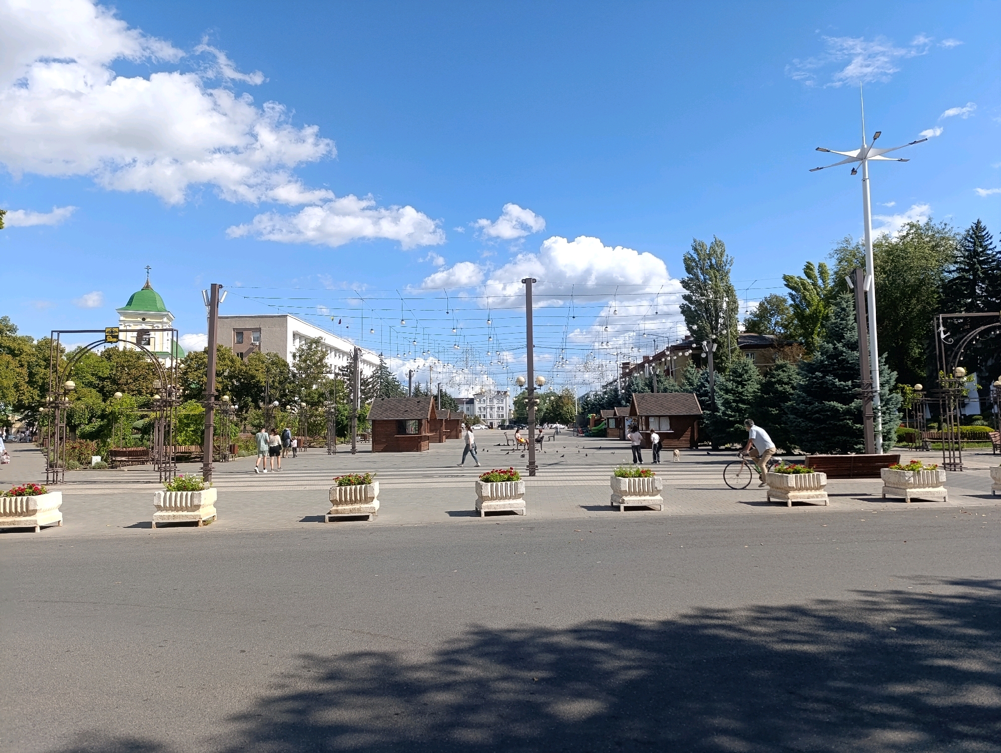 The image depicts a view of a city square, likely in Bender, Moldova, on a sunny day with a clear blue sky dotted with white clouds. In the foreground, a paved road occupies the lower half of the frame, with shadows from unseen trees falling across it. Several large, rectangular stone planters filled with red flowers are positioned along the edge of the road.
Beyond the planters, a wide pedestrian area stretches into the distance, paved with light-colored stones. Several small, wooden market stalls with brown roofs are set up along the sides of this area. A variety of trees, including tall evergreens and leafy deciduous trees, line the pedestrian zone and the background.
In the background, a building with a green domed roof and a cross, possibly a church or cathedral, is visible to the left. Further down the pedestrian area, there are several people walking, and one person is riding a bicycle. A tall, modern streetlamp with multiple light fixtures stands prominently on the right side of the square. String lights are suspended across the pedestrian area, connecting poles and adding to the ambiance. The overall scene suggests a bustling public space in a city.