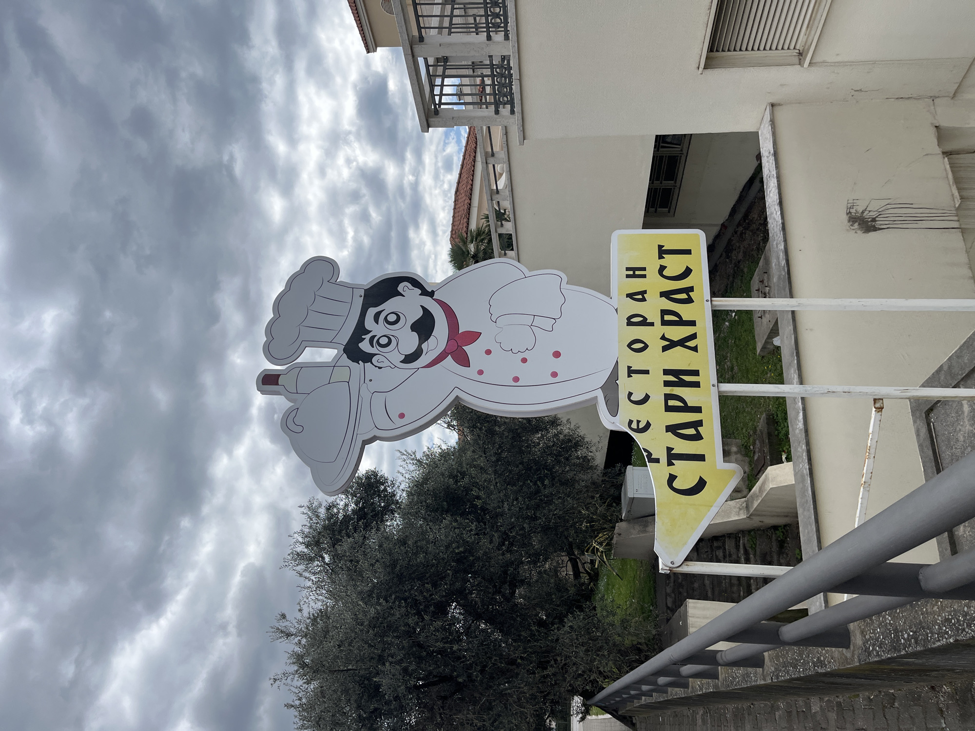 The image shows a sign for the "STARI HRAST" restaurant in Boreti, Montenegro. The sign features a cartoon chef holding a wine bottle and a plate. The sign is yellow with black lettering and points to the right. The sign is placed near a gray metal railing and a tree. In the background, there is a building with a balcony. The sky is overcast.
