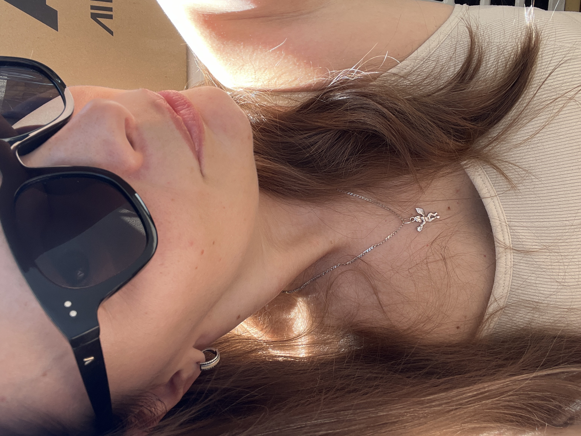 A close-up, profile shot captures the upper body of a fair-skinned woman wearing large, black sunglasses. She has light brown hair and is wearing a silver pendant on a delicate chain, featuring a small winged figure. A subtle hoop earring is visible on her left ear. Her attire appears to be a ribbed, light-colored tank top. The lighting suggests an outdoor setting, with sunlight creating shadows and highlights on her skin and hair. In the background, a brown cardboard box with the black lettering "AIL" is partially visible. The location is Tolyatti, Russia.