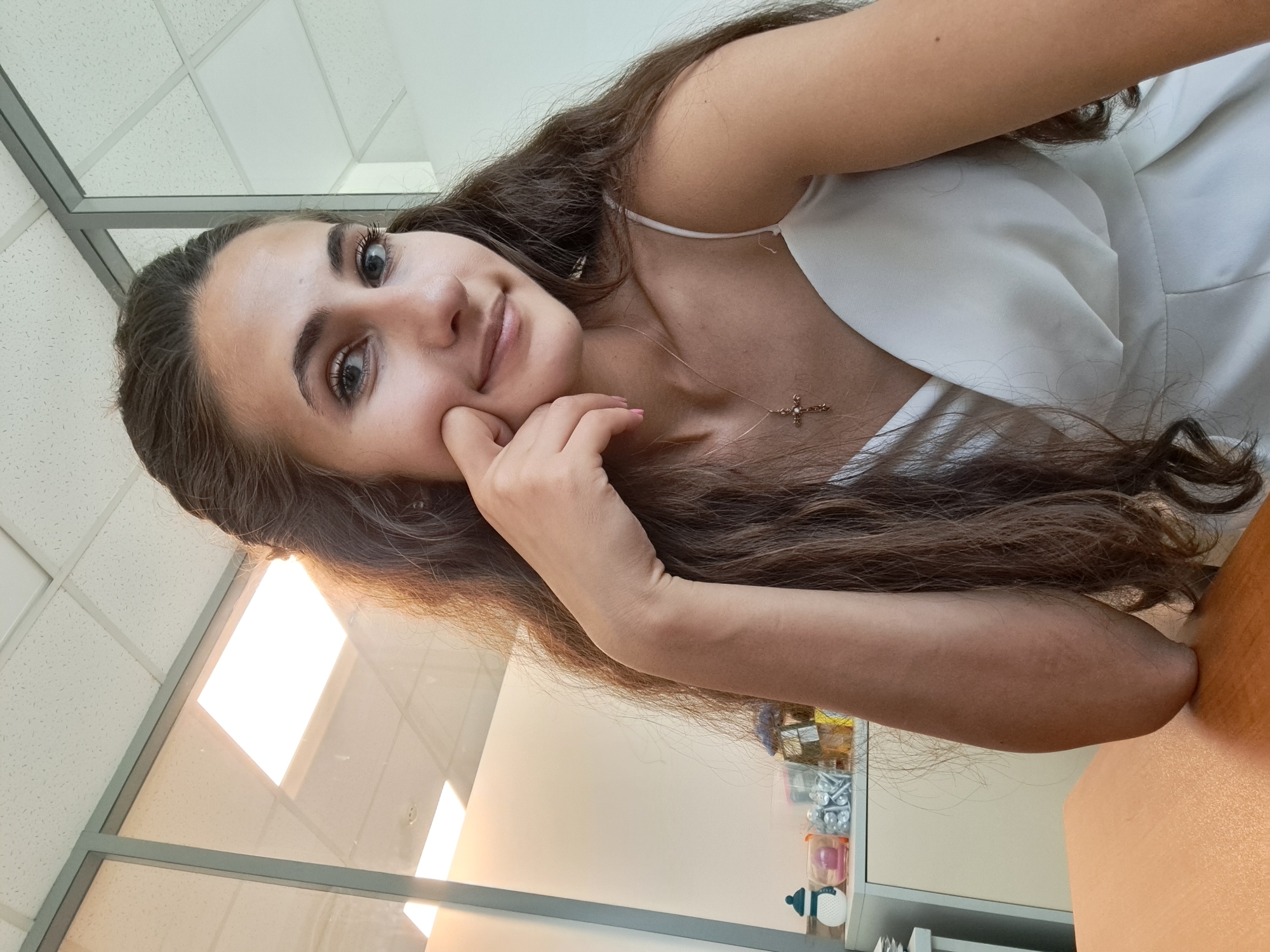 A young woman with long, dark brown hair is seated at a wooden desk in an office. She is looking at the camera with a slight smile, her right hand resting on her cheek. She is wearing a white, sleeveless dress with a V-neckline and a delicate gold necklace with a cross pendant. The background shows office partitions made of glass and metal framing, with ceiling lights and a white wall. A cabinet with various containers is visible on the left side of the image. The overall lighting suggests an indoor environment during daylight hours.