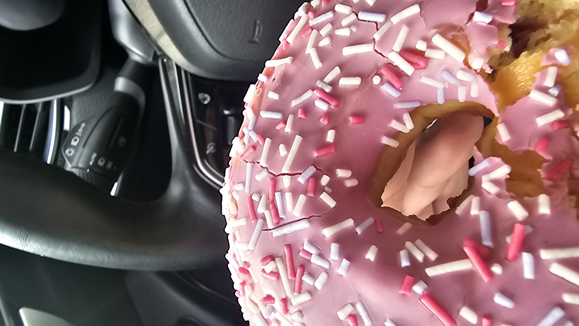 A close-up, vertical shot shows a pink frosted donut with white and pink sprinkles. The donut has cracks in the frosting and appears to be partially eaten. In the background, the steering wheel and dashboard of a car are visible, suggesting the donut is in a vehicle. The steering wheel has controls for lights and wipers. This image was taken in Kamensk-Uralsky, Russia.