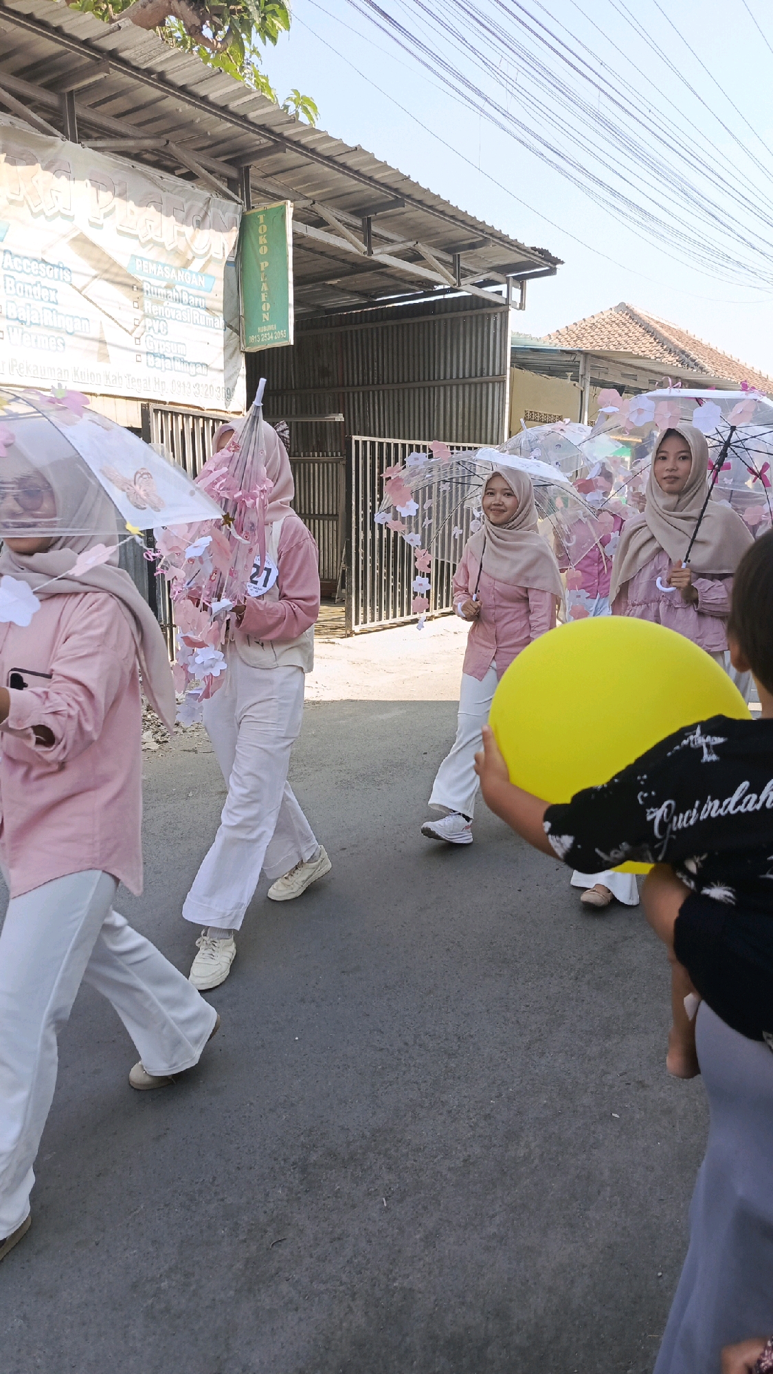 In Pekauman Kulon, Indonesia, a group of young women, likely participating in a parade or event, walk down a paved street. They are dressed in matching light pink long-sleeved shirts and white pants, with some wearing hijabs in a neutral tone. Each woman carries a clear umbrella decorated with paper flowers and butterflies. One woman wears a bib with the number '21' on it. In the foreground, a person's arm holds a large yellow balloon. The background shows a street with corrugated metal buildings and a tiled roof, suggesting a commercial or residential area. The sky is clear and bright, indicating daytime.