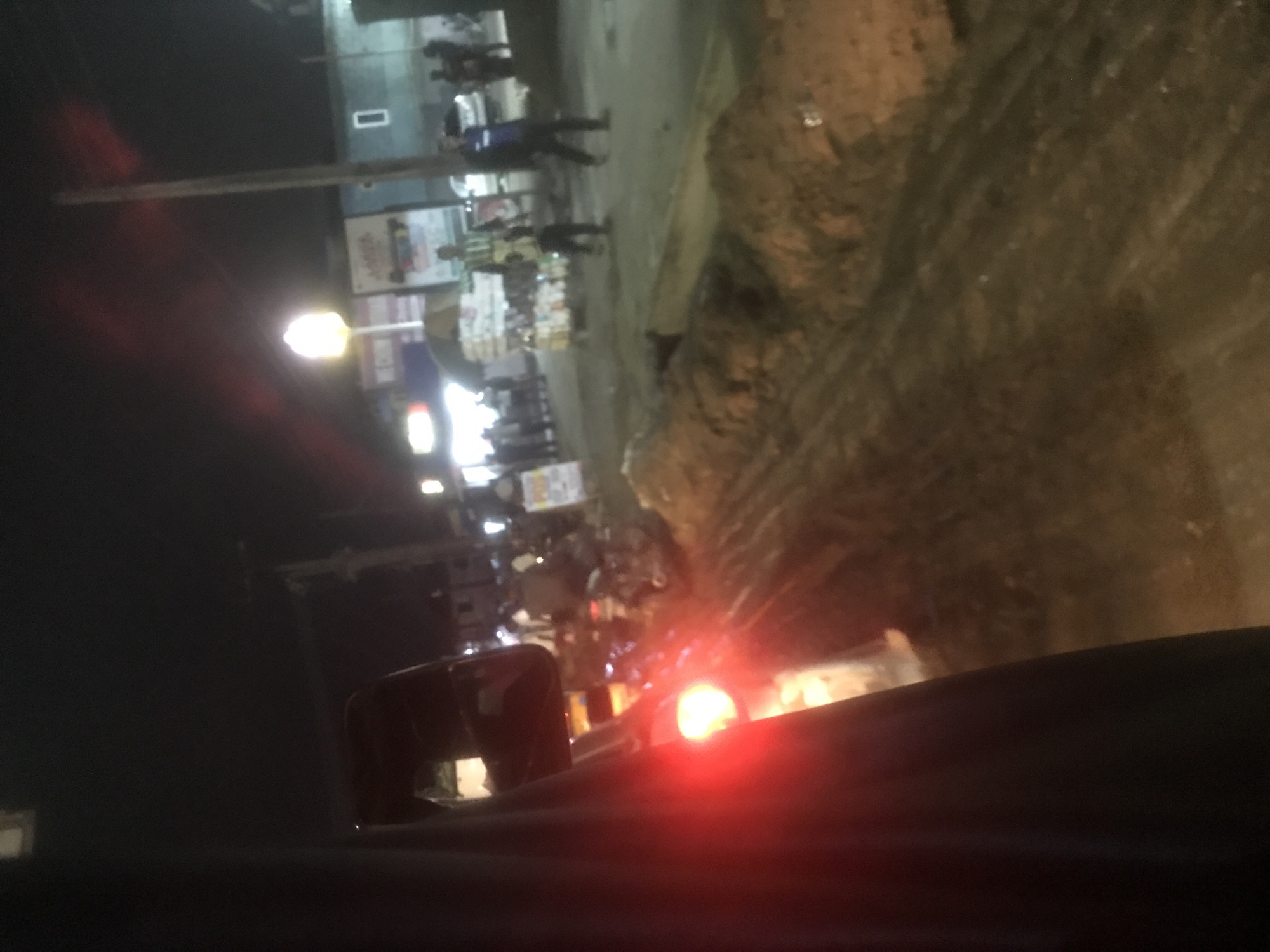 The image depicts a street scene at night in an unspecified urban location. A portion of a dark vehicle occupies the left side of the frame, with a red taillight visible and illuminated. The street appears to be unpaved and muddy, with piles of dirt and debris. Several individuals are present, some standing and some walking. A light pole with a glowing lamp stands prominently in the background, and signage is visible on buildings. The overall impression is of a busy, dimly lit street with ongoing activity.