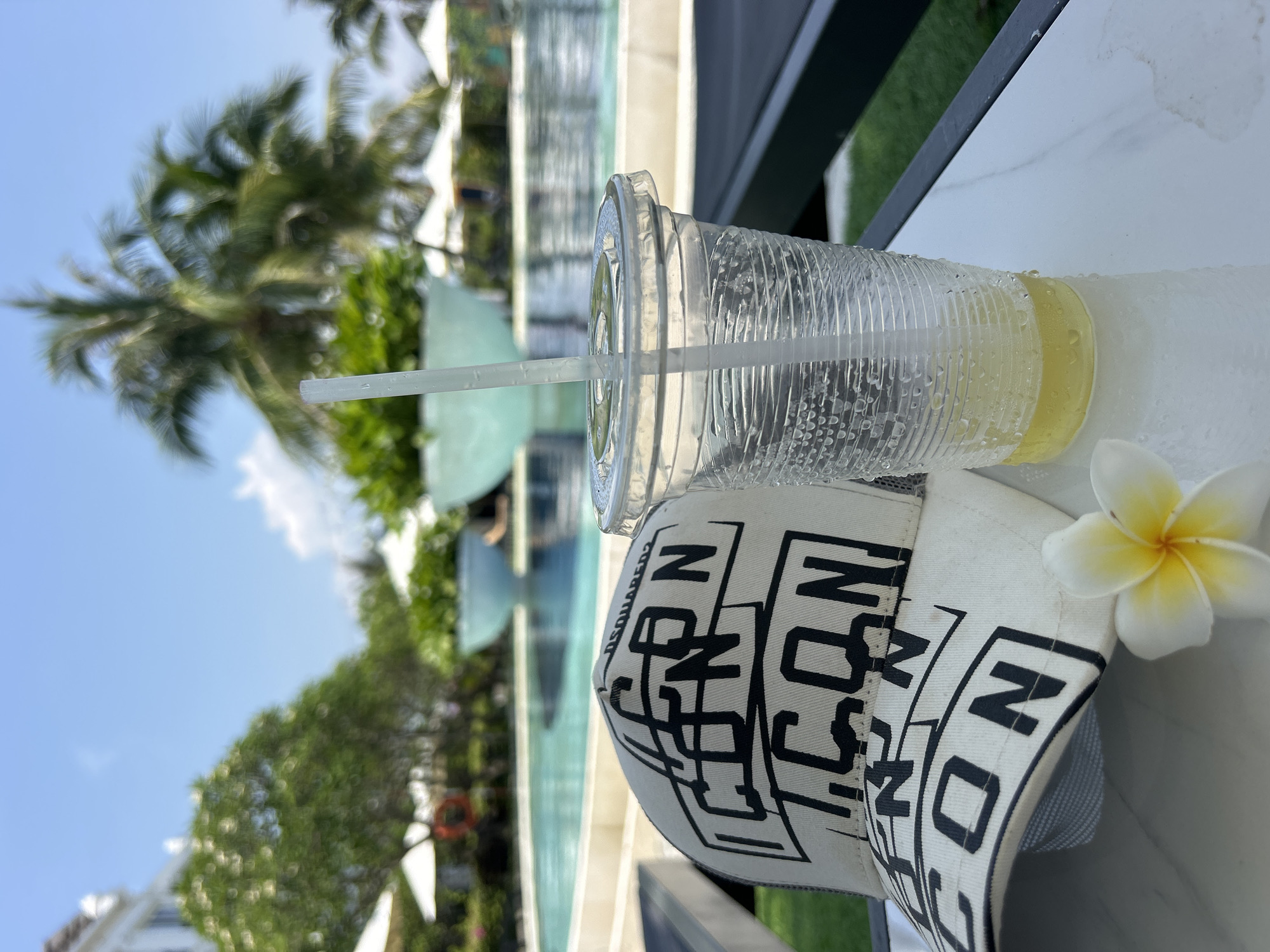 The image depicts a clear plastic cup with a straw, partially filled with a yellowish liquid. The cup is resting on a white baseball cap with the word "ICON" printed repeatedly in bold black letters. Next to the hat is a white and yellow plumeria flower. The scene is outdoors, likely near a resort or pool area, with a palm tree, foliage, and a body of water visible in the background. The background suggests that the location is in Bac Nha Trang Ward, Vietnam, as it is known for such features.
