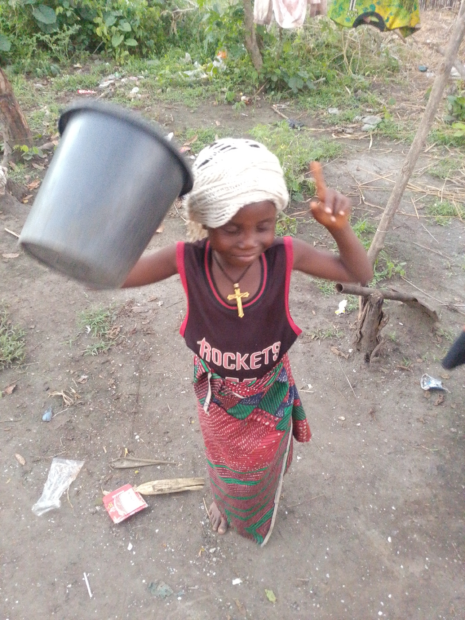 A young girl stands in a dirt yard in Otulu, Nigeria, holding a large grey bucket in her left hand. She wears a white headscarf, a dark sleeveless shirt with red trim that says "ROCKETS" in white letters and a gold cross necklace, and a colorful red, green, and black patterned skirt. She is looking down and to her right. Her right index finger is pointed upwards and slightly to her left. In the foreground, on the ground, are various pieces of debris, including a plastic bag, a red and white packet, and some dried leaves and twigs. The background consists of greenery and some wooden structures.