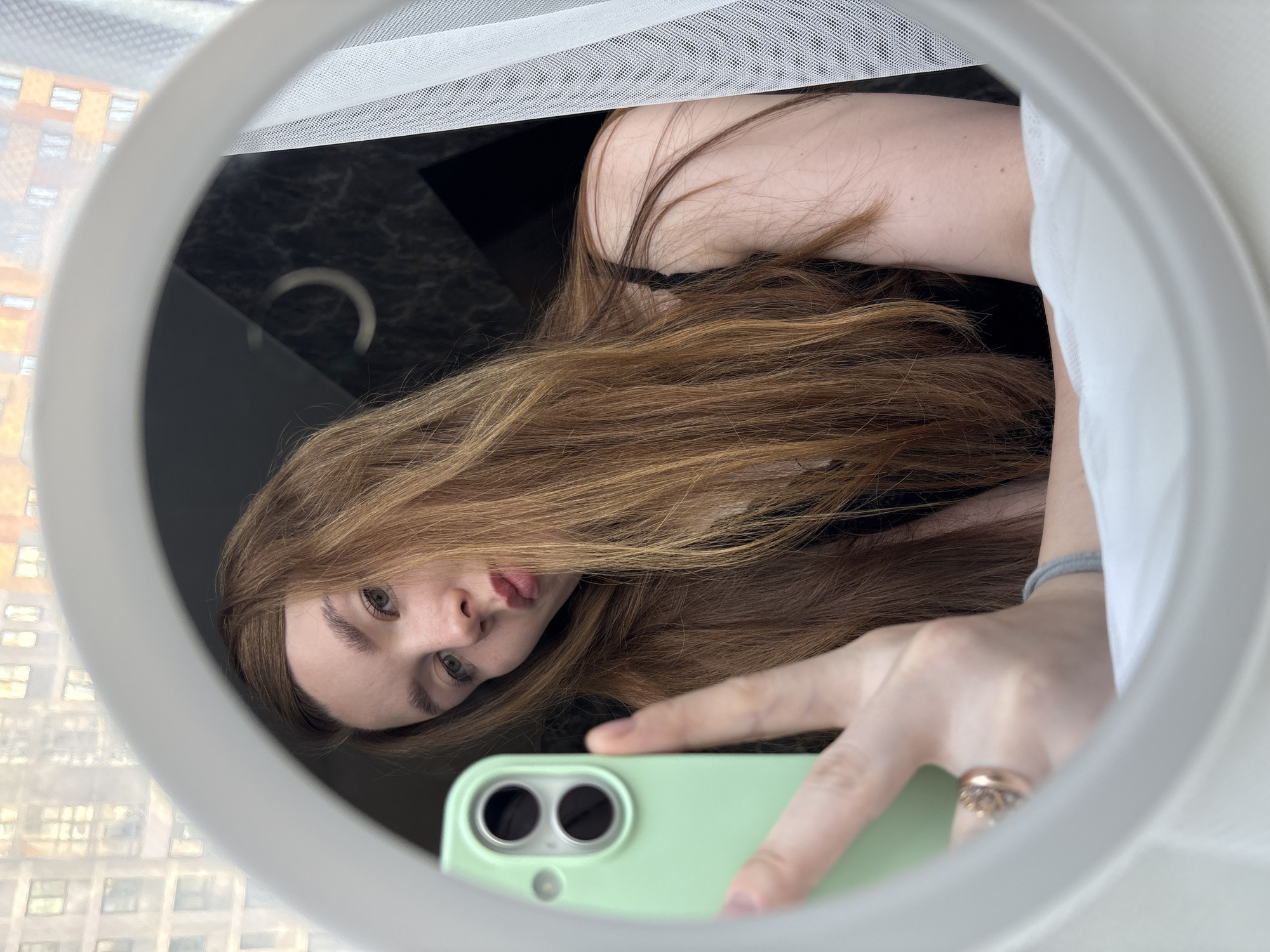 The media file is a selfie taken in a mirror, with the phone held by the subject's hand. The subject is a person with long, light brown hair, and appears to be indoors. The reflection in the mirror shows the person's face, looking directly at the camera. The person is wearing a black top and a ring on their finger. Behind the subject, a white sheer curtain is visible, and beyond that, a blurred view of buildings, suggesting an urban setting, possibly from a window. The lighting appears to be natural, indicating it could be daytime. The overall mood of the image is casual and personal.