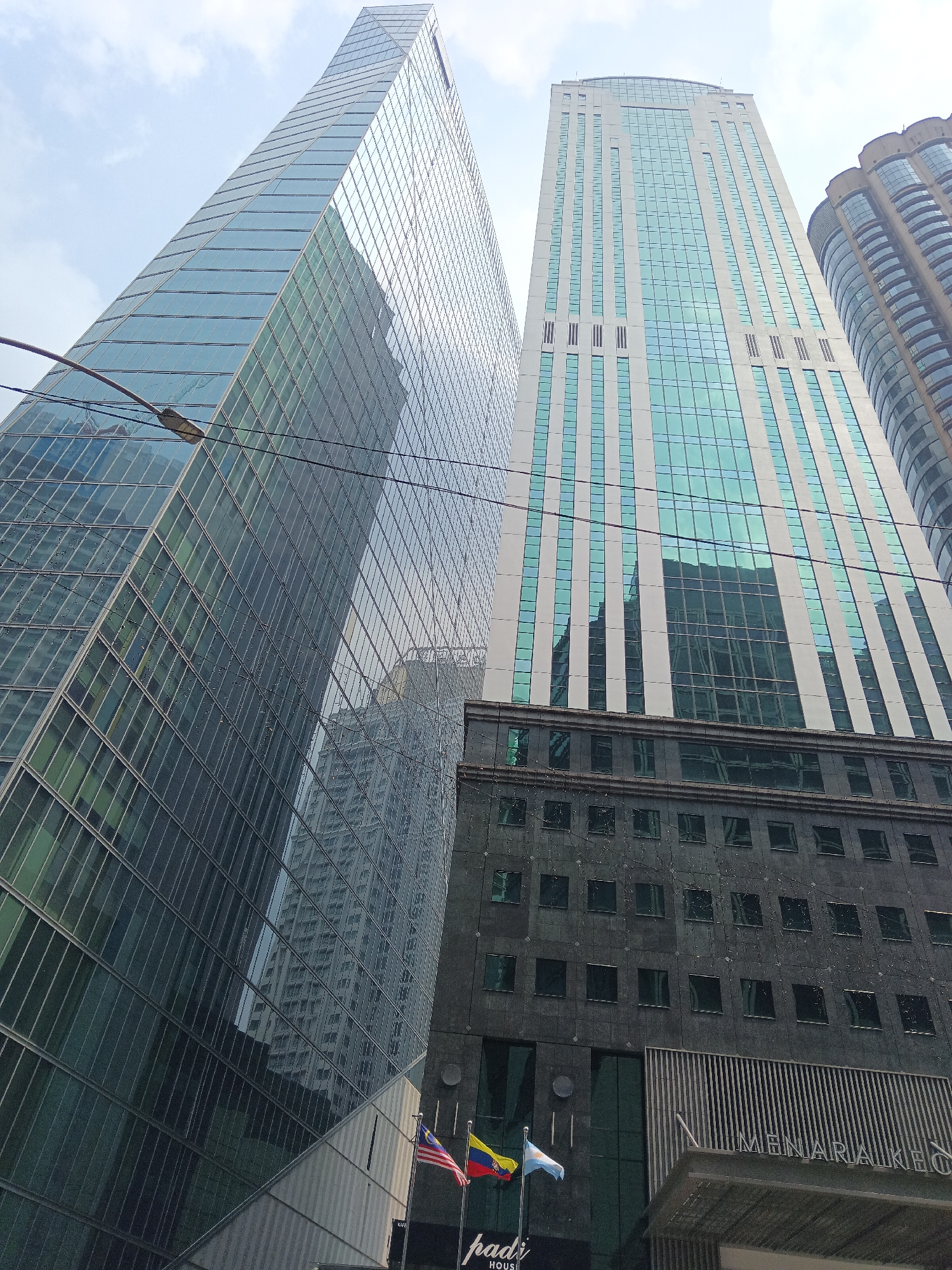 The image shows a low-angle view of modern skyscrapers in Kuala Lumpur, Malaysia. The foreground features a building with a dark, textured facade and numerous dark-tinted windows. To the left, a tall, glass-facade skyscraper dominates the view, its surface reflecting the sky and other buildings. To the right of this glass skyscraper is another tall building with a lighter facade featuring vertical elements and rows of turquoise-tinted windows. A portion of a third, curved building with a lighter facade and rounded windows is visible on the far right. In front of the dark-facade building, two flags are displayed: the Malaysian flag (Jalur Gemilang) and what appears to be the flag of Colombia. Text on the building below the flags reads "MENARA KEO" and partially visible "padi" with "HOUS" below. The sky is overcast with scattered clouds. Overhead power lines are visible in the upper portion of the image.