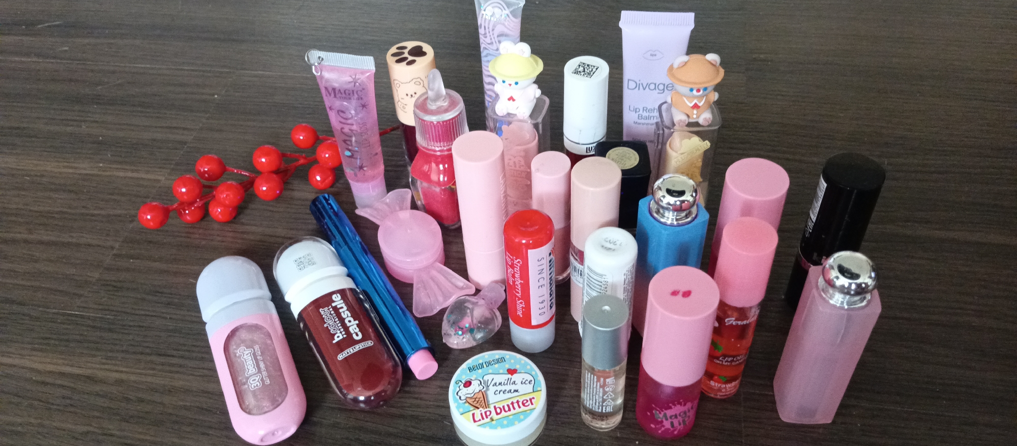 The image shows a collection of various lip care and cosmetic products arranged on a dark brown wooden surface. The items include lip glosses, lip balms, lipsticks, and lip butters in a variety of packaging, colors, and sizes. Some of the brands and product names visible are "Magic Lip Gloss," "B.colour Capsule Matte Lipstick," "3Q Beauty Glitter Shiny Lip Gloss," "Taftaia Strawberry Shine Lip Balm," "Belor Design Vanilla Ice Cream Lip Butter," and "Divage Lip Rehab Balm Marshmallow." Some of the lipsticks are in traditional tubes, while others are in squeeze tubes or jars. There is also a decorative element of red berries on a branch.