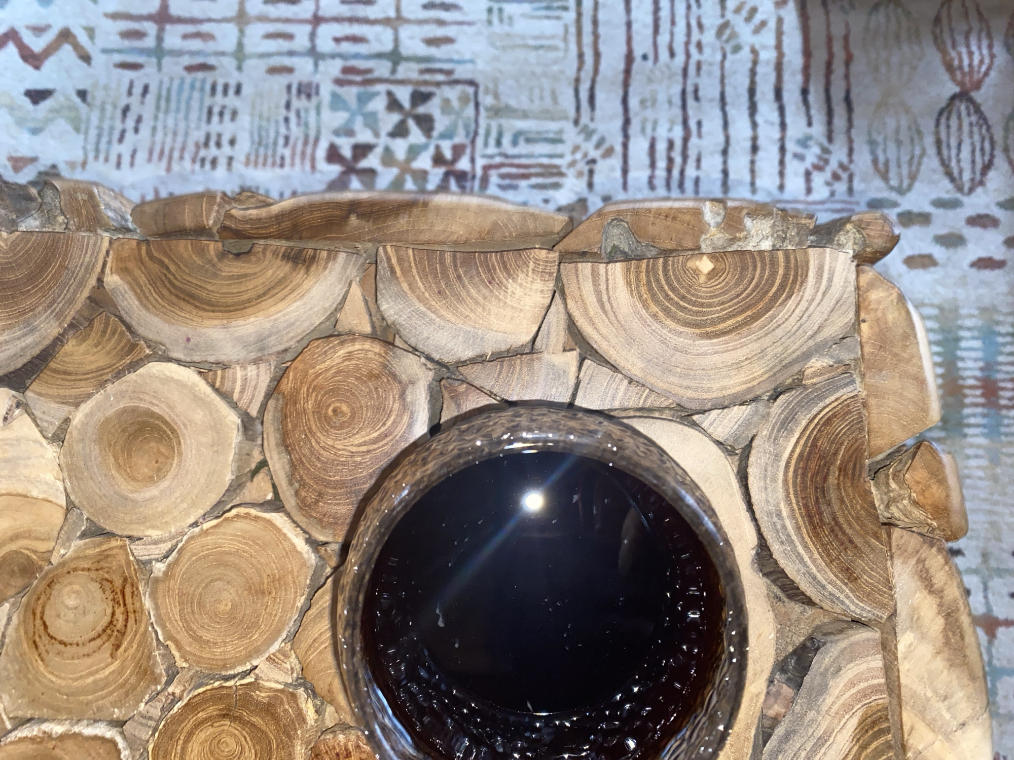 The image shows a close-up of a glass of dark liquid, possibly coffee or tea, resting on a surface made of circular wooden cross-sections. The wooden pieces are arranged together to form a textured surface. To the right of the wooden surface is a patterned fabric, possibly a tablecloth or a rug. The lighting in the image suggests an indoor setting. The scene is static, with no visible people or actions taking place. The location of this scene within Krasnoflotskoe, Russia, is not specified by the image itself.