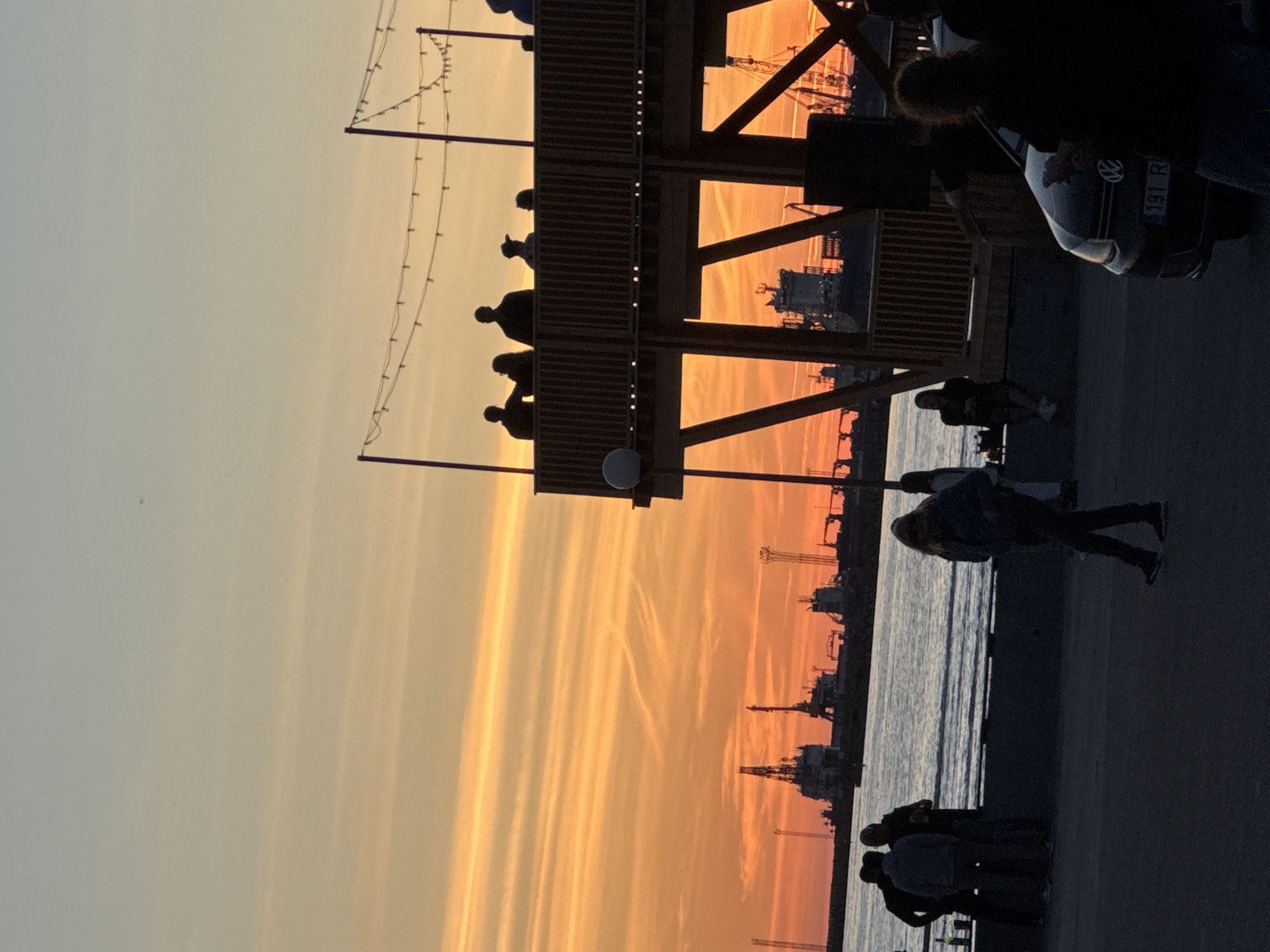 The image captures a sunset scene at the Tallinn Seaplane Harbour, Estonia, with a vibrant orange and yellow sky. In the foreground, silhouetted figures of people are visible on a promenade and on a wooden structure. The wooden structure, which appears to be an elevated deck or cafe, has string lights draped across it and a railing along its edge. Several people are standing or sitting on this structure, also silhouetted against the sunset.
Further in the background, across a body of water, is a harbor with various industrial structures, including cranes and large ships, silhouetted against the colorful sky. The water reflects the sky's colors. On the promenade in the foreground, a group of people are standing and talking near the water's edge. Another person is walking away from the viewer. A car with a license plate "191 R" is parked on the right side of the frame, partially in silhouette. The overall atmosphere suggests a casual gathering or event by the waterfront during twilight.