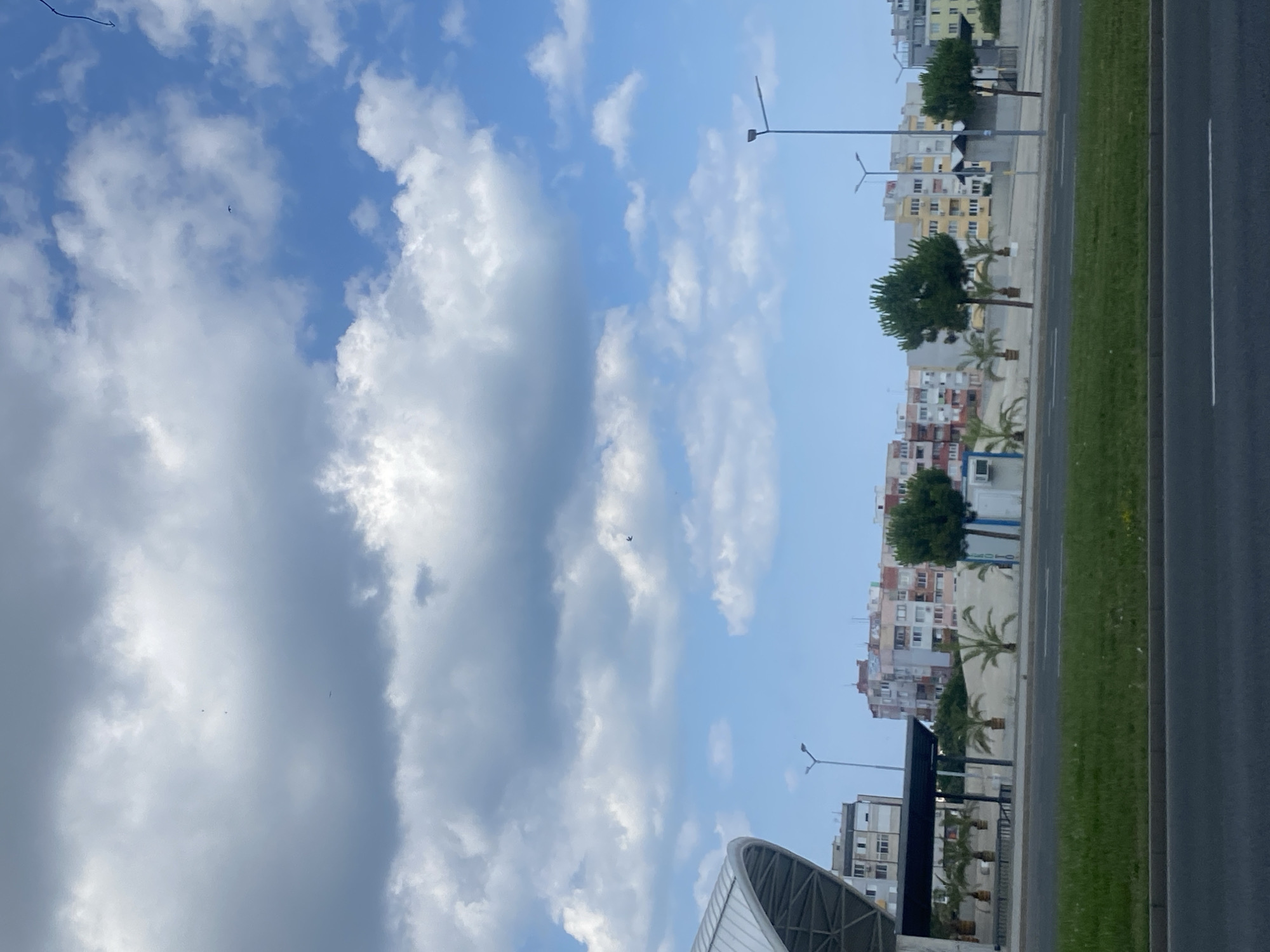 The image captures a street scene in Burgas, Bulgaria, likely during daylight hours with scattered clouds. In the foreground, a dark asphalt road with white markings is visible, bordered by a strip of green grass. Beyond the grass, a light-colored concrete wall runs horizontally, separating the road from a paved area with several palm trees. A modern, curved white roof structure, possibly part of a public transport stop or building, is visible on the far left, extending over a covered seating area. In the middle ground, a row of multi-story buildings with colorful facades, characteristic of urban residential areas, stretches across the horizon. Several streetlights are also present. The sky is a mix of bright blue and gray clouds, with a few small objects, possibly birds or distant aircraft, visible in the sky. No people are clearly identifiable in the image.