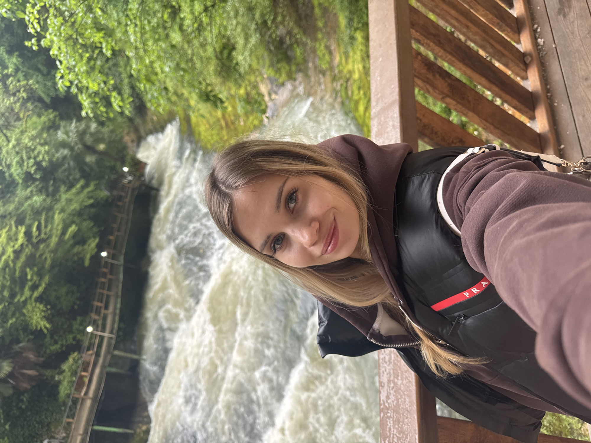 A young woman is positioned on a wooden walkway alongside a waterfall in Tsabal, Abkhazia. She is wearing a dark brown hoodie with a black Prada vest and a light-colored shoulder bag. She has blond hair and light skin. The waterfall is behind her, with white, rushing water flowing down from the top. Green foliage is visible in the background. The wooden walkway includes a handrail made of wooden beams. The image appears to be a selfie.