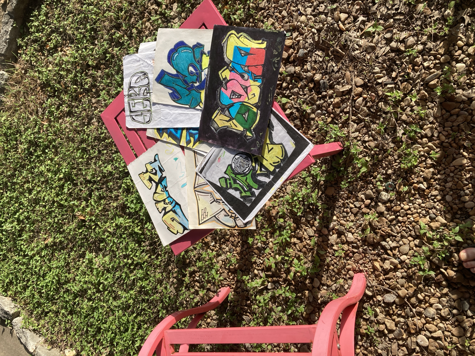 A collection of graffiti-style drawings is displayed on a pink chair outdoors. The drawings are on various types of paper and feature colorful, abstract lettering. They are arranged in a pile on top of the chair. The surrounding environment consists of green grass and gravel. The image is taken from a high angle, looking down at the chair and drawings.