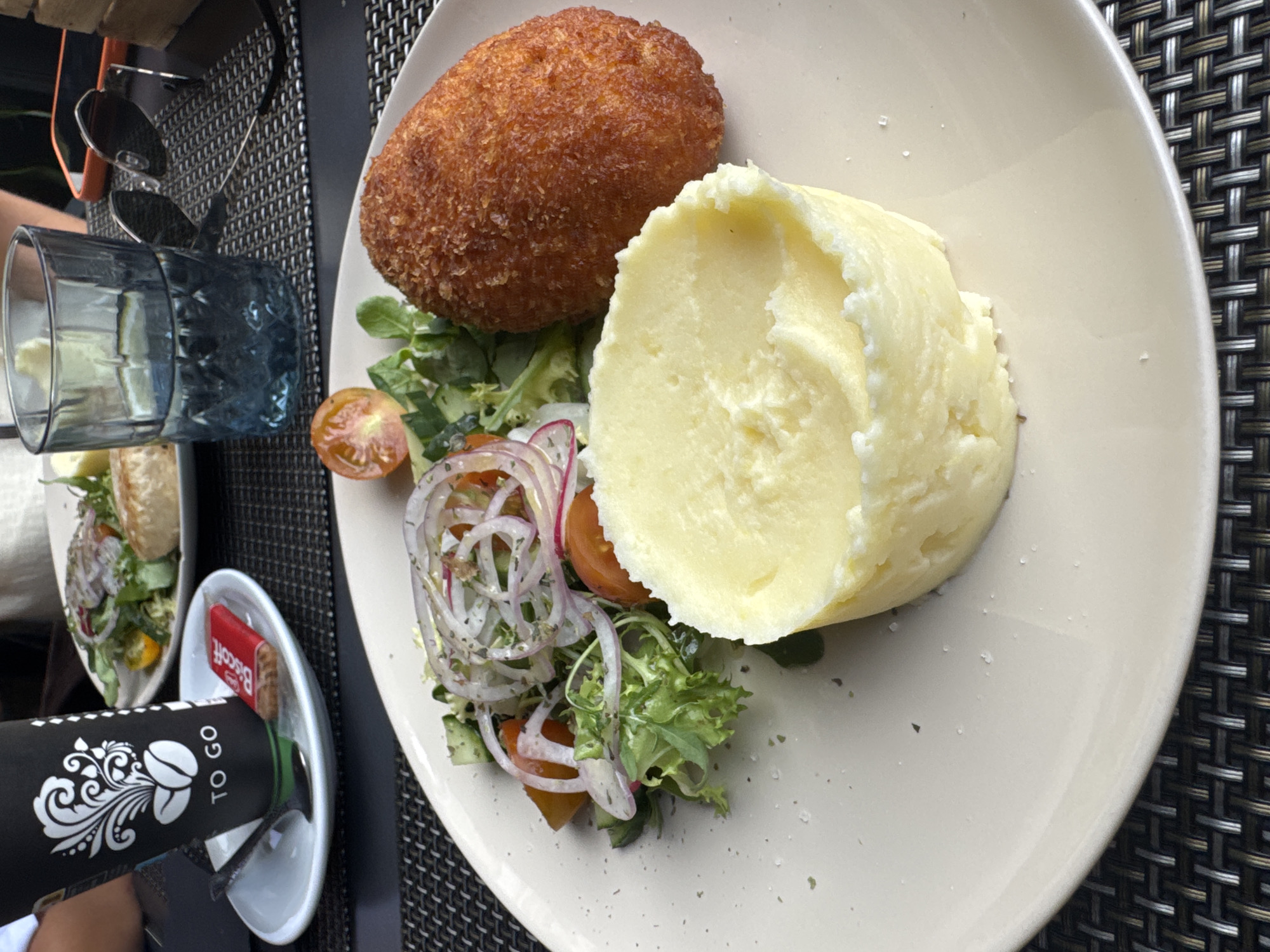 This image shows a close-up view of a meal served on a white plate, set against a dark, woven placemat. The meal consists of a golden-brown, breaded cutlet, a generous portion of mashed potatoes, and a small side salad. The salad includes mixed greens, sliced red onion, and halved cherry tomatoes.
To the left of the plate, a blue glass filled with a clear liquid is visible. In the foreground, a black "TO GO" coffee cup with white decorative elements and coffee bean imagery is placed in a small white dish. This dish also holds a small red packet labeled "Biscott" with a visible biscuit inside. Sunglasses rest on the placemat near the glass.
The lighting suggests it is daytime. The overall setting appears to be an outdoor dining area, possibly a restaurant patio, given the presence of a placemat and tableware. The meal is presented neatly, indicating a dining experience. The image is captured from a top-down perspective, focusing on the food and immediate surroundings. No people are clearly visible, though a hand may be partially present near the coffee cup. The meal appears to be a hearty dish, suitable for lunch or dinner. The environment seems casual and relaxed.