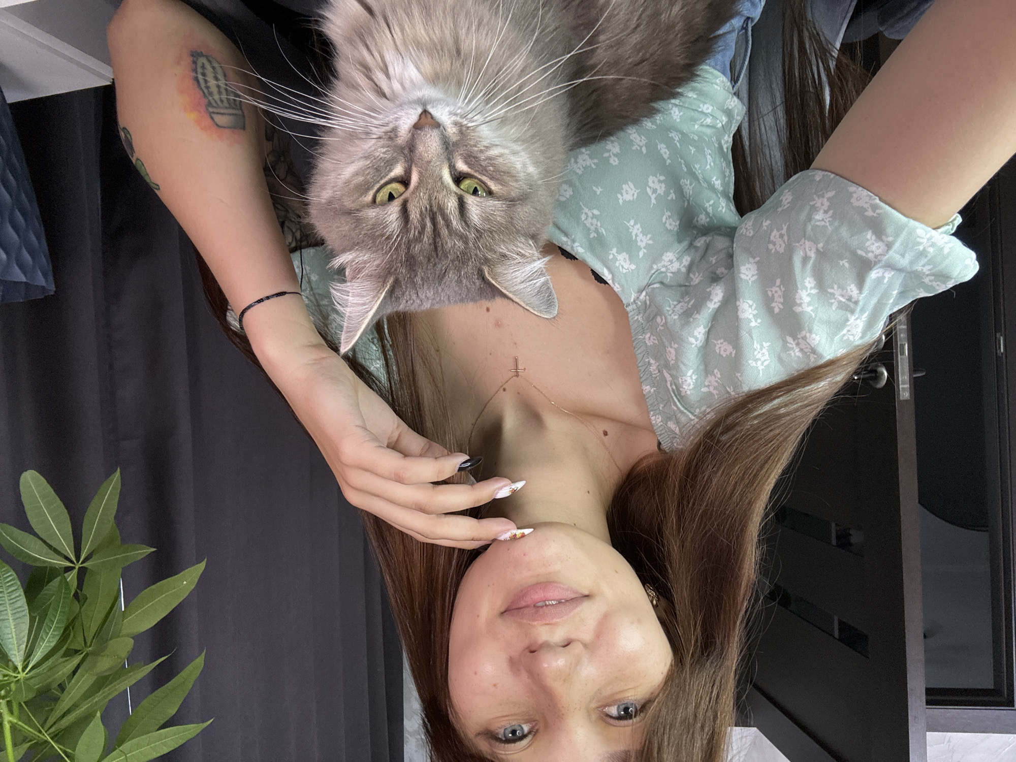 A young woman with long brown hair and blue eyes is sitting with a grey cat on her lap. She is wearing a light green floral print top and a necklace with a cross pendant. She is looking at the camera and smiling slightly, with her hand resting on her chin. The cat is looking at the camera with its green eyes. In the background, there is a dark curtain, a plant, and a doorway. The scene appears to be indoors, in a home environment. The image does not contain information that would allow for its location to be specified within the city of Novokuznetsk, Russia.