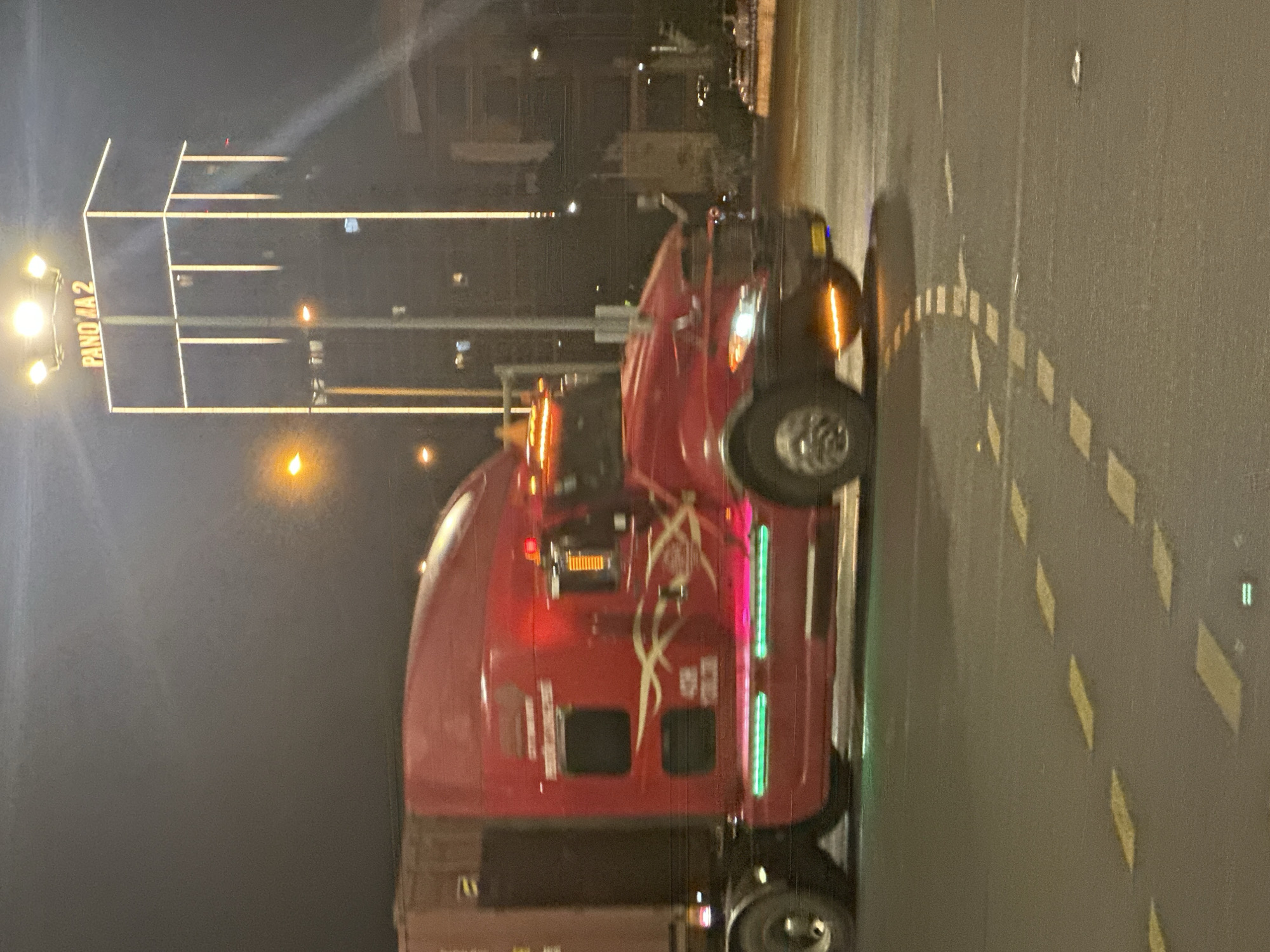 A red semi-truck with green undercarriage lights drives on a city street at night. The truck has a trailer attached. A tall building with "PANO 2" lit up on top is in the background. Streetlights and building lights illuminate the scene. The road has yellow lane markings. The image appears to be taken in Da Nang, Vietnam, judging by the building's signage and the overall urban aesthetic.