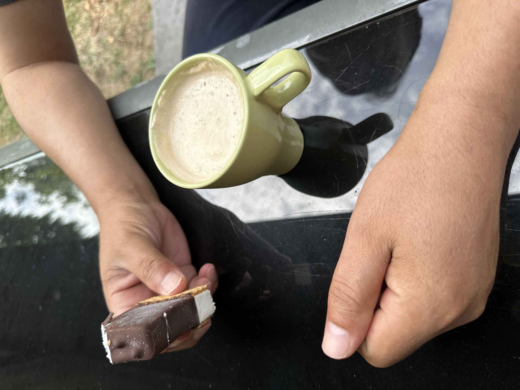 The image shows a close-up of two hands and a light green mug of coffee on a dark, reflective surface, likely a table. One hand, in the upper right, is holding a portion of a chocolate-covered ice cream bar that appears to have a white layer beneath the chocolate and some shredded coconut on top. The other hand, in the lower left, is resting with fingers slightly curled. The mug is filled with a frothy coffee beverage. The background is out of focus, showing greenery and what appears to be an outdoor setting. The image does not provide any specific visual cues that would allow for the determination of its location within Krefeld, Germany.