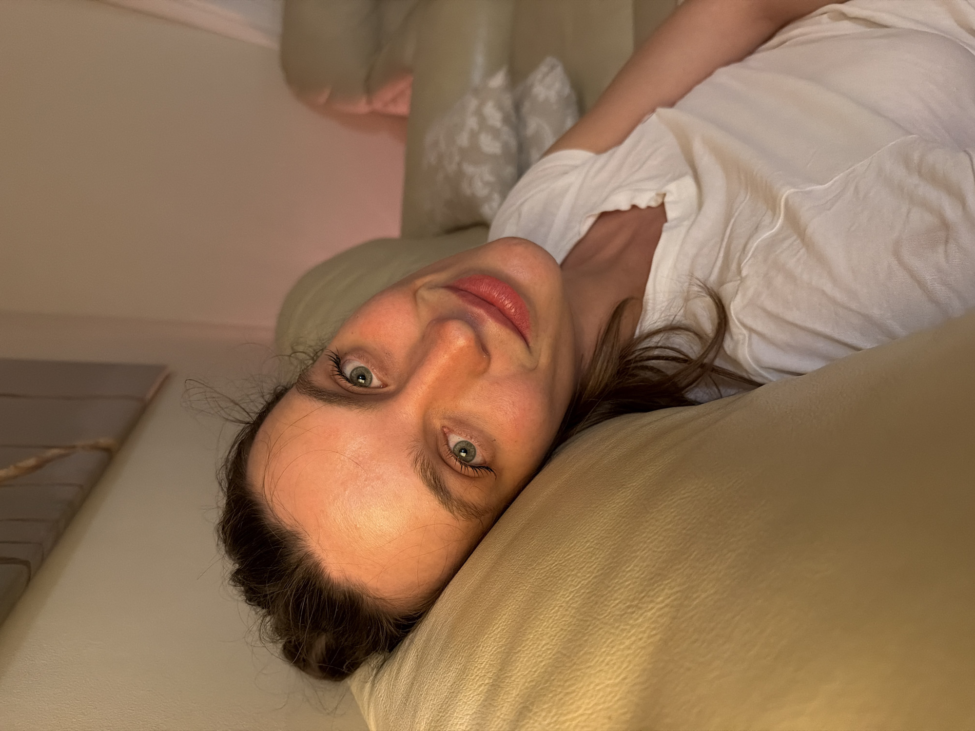 A woman with fair skin, dark hair, and striking light green eyes is captured in an indoor setting. She is lying on her side, resting her head on a light beige or cream-colored couch or bed. Her dark hair is casually styled around her face. Her lips are slightly parted, revealing a subtle, warm-toned lipstick. She is wearing a simple white short-sleeved top. Her gaze is directed straight at the camera with a calm, inviting expression and a hint of a smile.
The lighting in the room is soft and warm, suggesting an evening or nighttime scene indoors. In the background, additional light-colored upholstered furniture with decorative pillows featuring a light pattern can be observed. The scene depicts a moment of relaxation or a casual portrait. There is no visible text in the image. While the location context is provided as Budva, Montenegro, no specific visual cues within the image confirm this.