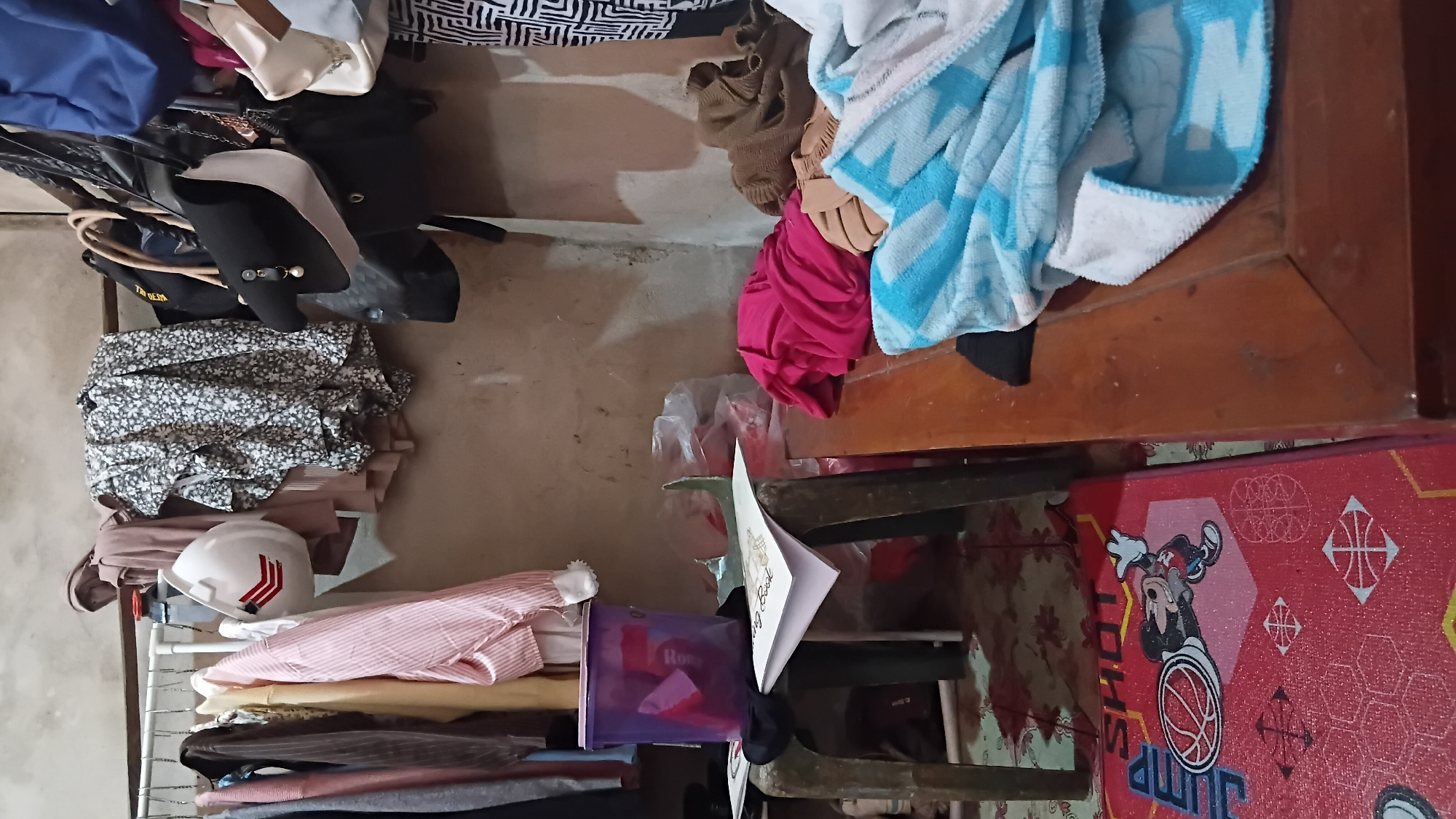The image depicts a cluttered domestic interior, likely a bedroom or storage area, in the city of Prabumulih, Indonesia. A clothes rack dominates the upper left and center, laden with various garments including shirts, dresses with floral patterns, and what appears to be a hard hat with red stripes. Several bags, including a black and white handbag and larger black bags, hang from a bar above the clothes. To the left, a small, dark green plastic stool supports a purple bucket filled with items and a spread-out book. In the foreground, a wooden surface, possibly a table or bench, is covered with folded textiles, including a bright pink garment and a blue and white striped towel. A red floor mat with a cartoon character and basketball motifs is visible on the lower left. The walls are unpainted and show signs of wear. The overall impression is one of a lived-in space with items not neatly organized.