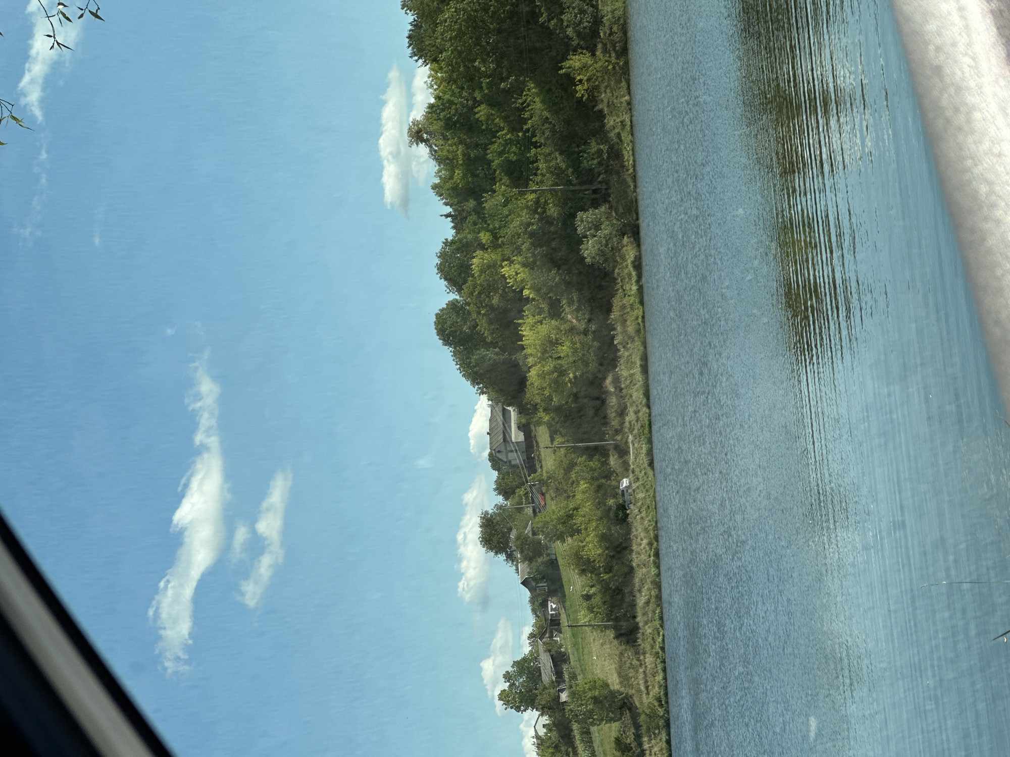 A wide shot captures a calm body of water in the foreground, with gentle ripples reflecting the sky and surrounding landscape. A green, grassy bank separates the water from a small village nestled on a hillside dotted with trees and houses. The houses appear to be of varying sizes and architectural styles, some with dark roofs and others with lighter ones. A white car is parked on a dirt road running along the edge of the village. The sky above is a clear blue, with a few fluffy white clouds scattered across it. The scene is illuminated by bright daylight, suggesting it is daytime.