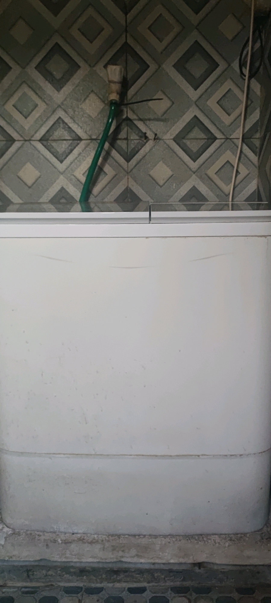 The image shows a white freezer with a light gray base, positioned against a tiled wall. The tiles are diamond-shaped in alternating shades of gray and light green. A green hose with a light-colored attachment, possibly a brush or nozzle, is coiled and secured to the wall with black zip ties. A white electrical cord runs vertically down the right side of the frame, also against the tiled wall. The floor in front of the freezer is tiled with a dark gray hexagonal pattern. The scene appears to be indoors, possibly in a utility area or kitchen, given the presence of the freezer and the tiled surfaces. The freezer is clean, with some minor scuff marks visible on its surface and base. The overall impression is one of a functional appliance in a common domestic or commercial setting in Gambarsari, Indonesia.