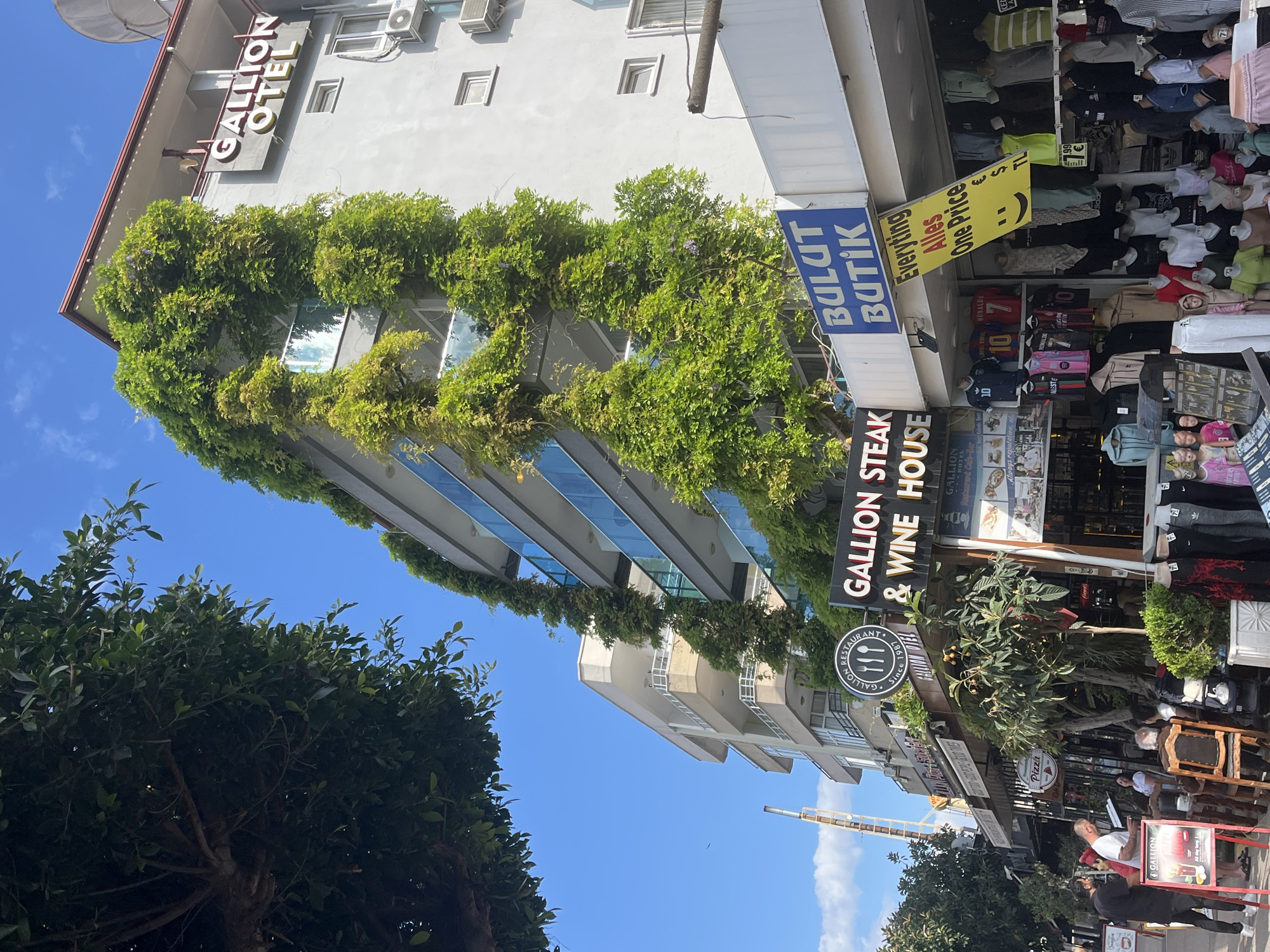 The image shows a street view in Alanya, Turkey. The building on the left, "GALLION OTEL", is covered with greenery. Adjacent to the hotel is "BULUT BUTİK", a clothing store with items on display outside. A yellow sign with a smiley face promotes "Everything Alles One Price". Further along the street is "GALLION STEAK & WINE HOUSE", with outdoor seating. People are visible along the sidewalk, including a person in a white shirt looking at a menu or document. A crane or other tall construction equipment is visible in the background against a blue sky with some clouds. Trees are visible on the side.