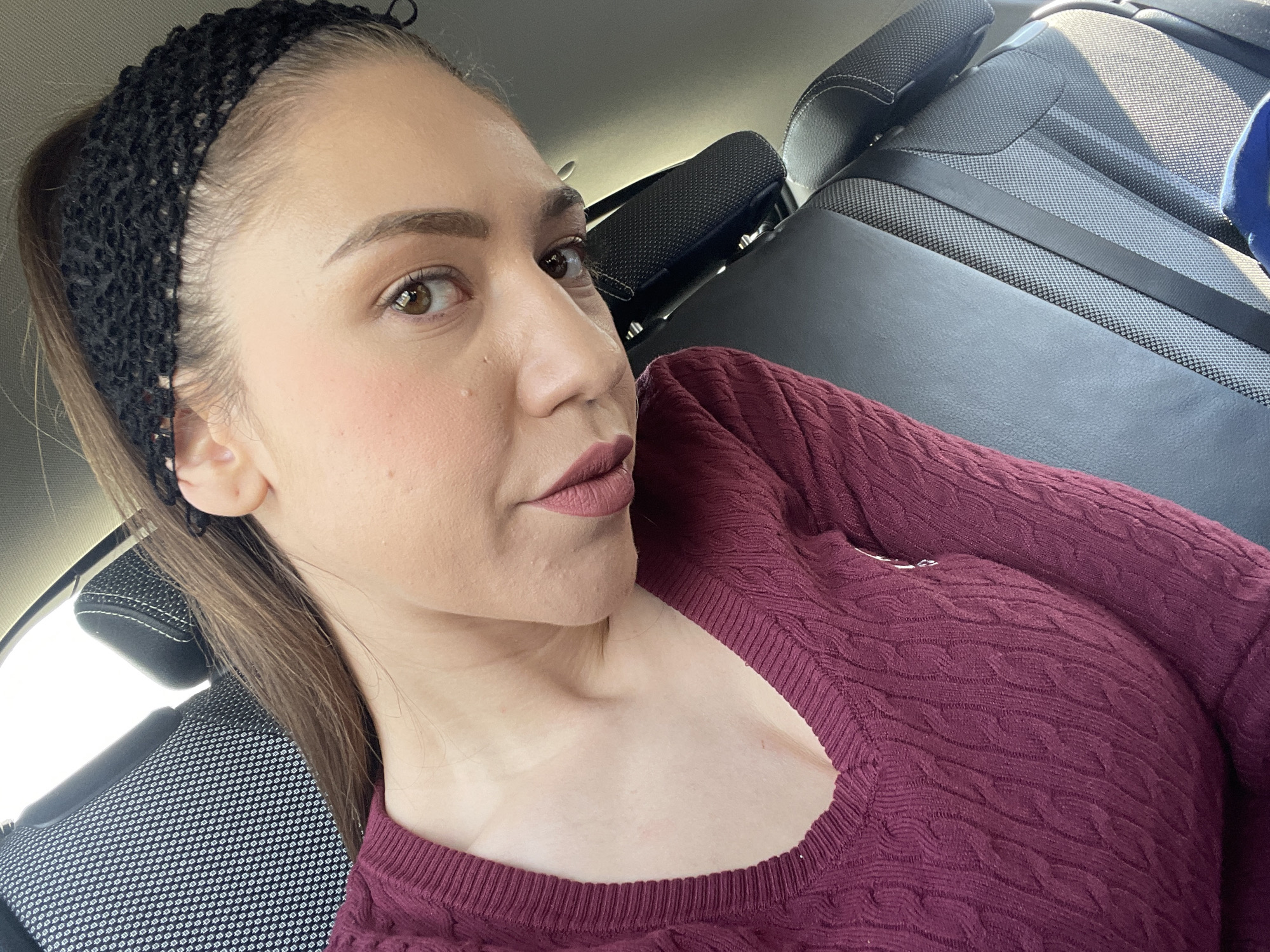 A woman is in the backseat of a vehicle. She has brown hair pulled back with a black headband, brown eyes, and is wearing a maroon cable-knit sweater and maroon lipstick. The car seats are grey with black headrests. The location is unspecified and is unrelated to Dobrogled, Bulgaria. There is no discernible information to indicate the city of Dobrogled, Bulgaria, or any activities or interactions in the described location.