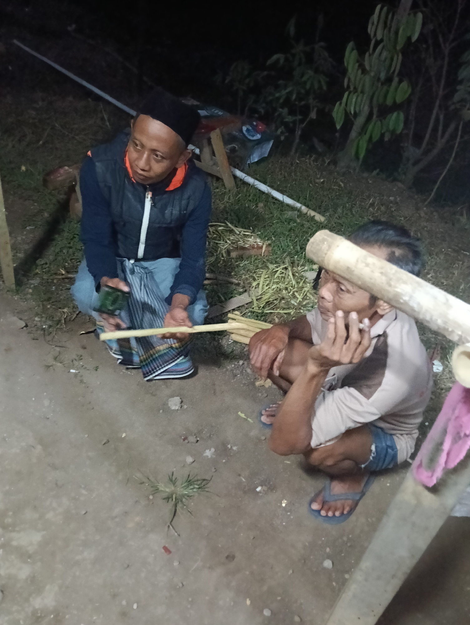 Two men are squatting outdoors at night. The man on the left is wearing a black peci hat, a dark vest over a blue shirt, and a sarong. He holds a green object in his left hand and a piece of bamboo in his right. The man on the right is wearing a light-colored collared shirt and shorts. He is holding a cigarette in his right hand and resting his chin on his left hand. They appear to be in a rural or semi-rural setting, with vegetation and dirt ground visible. A white pipe is visible in the background. The lighting suggests artificial illumination. There is no discernible indication of a specific location within Tasikmalaya, Indonesia.