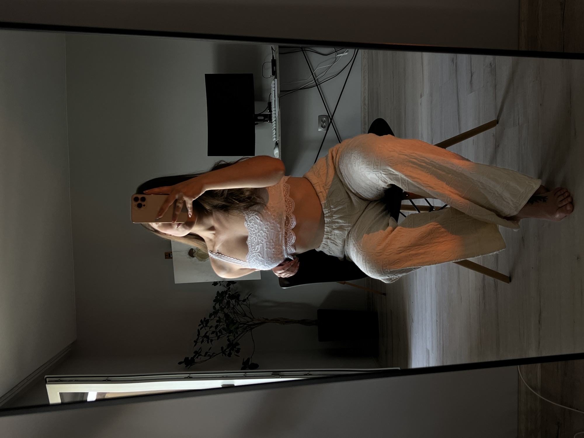 This media file is a mirror selfie taken indoors, presumably in a home office or living space. The primary subject is a young woman with long, dark hair, reflected in a large, floor-to-ceiling mirror. She is wearing a white lace bralette and loose-fitting, cream-colored pants. She is sitting on a chair, holding a smartphone to take the picture.
In the background, visible through the reflection, is a desk with a computer monitor and keyboard, suggesting a workspace. There are also electrical cords and a wall outlet. The flooring appears to be light-colored wood planks. A large potted plant with dark green leaves is also visible to the left of the woman. The lighting suggests it is either late afternoon or early evening, with warm light casting a glow on the woman's pants. The overall mood is casual and relaxed. There is no visible text in the image.
