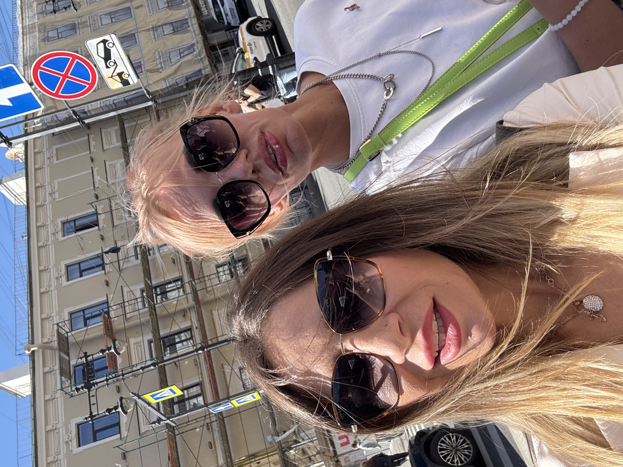 Two women take a selfie on a sunny day in Saint Petersburg, Russia. The woman on the left has long blonde hair and is wearing sunglasses and a light-colored jacket. The woman on the right has short blonde hair and is wearing sunglasses, a white t-shirt, and a bright green strap across her chest. Behind them, a building is undergoing renovation, with scaffolding visible. Traffic signs are also present, including a no-parking sign and a tow-away sign. A yellow taxi is visible in the background.