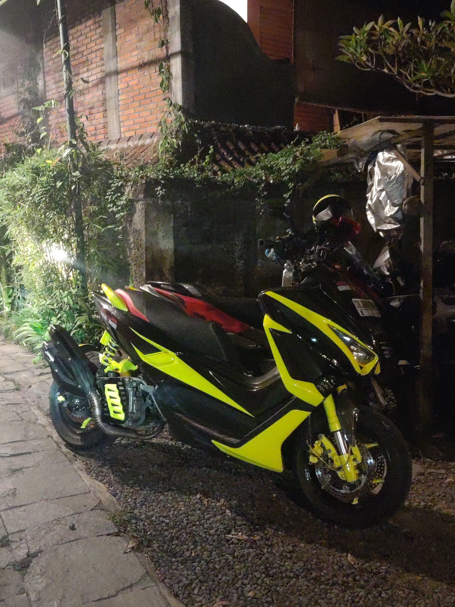 The image captures a custom-painted scooter parked outdoors at night in Ubud District, Indonesia. The primary subject is a large scooter, likely a maxi-scooter, featuring a striking two-tone design of black and neon yellow/lime green. The vibrant yellow is prominently displayed on the front fairing, side body panels, front suspension forks, brake calipers, rear shock absorbers, and elements around the exhaust system, suggesting significant customization. A black seat with subtle red highlights is visible, and a yellow and black helmet hangs from the handlebars.
The scooter is parked on a patch of gravel, next to a paved stone pathway that extends from the lower left towards the middle of the frame. In the background, a building or wall constructed from reddish-brown bricks is partially obscured by lush green climbing vines and foliage. Denser green plants, including bamboo-like stalks, grow on the left side, illuminated by a light source off-camera. On the right, parts of another dark-colored scooter and a silver, reflective cover are visible beneath an overhead structure, possibly a shelter or roof extension. Leafy tree branches are seen in the upper right.
The scene is set at night, with artificial lighting creating bright highlights on the scooter's vibrant yellow surfaces and illuminating parts of the surrounding vegetation, contrasting with the deep shadows. No human activity is depicted. The combination of brick architecture, traditional-looking stone pathways, and abundant tropical foliage aligns with the specified location context of Ubud, Indonesia. No discernible text is legible on any objects in the image.