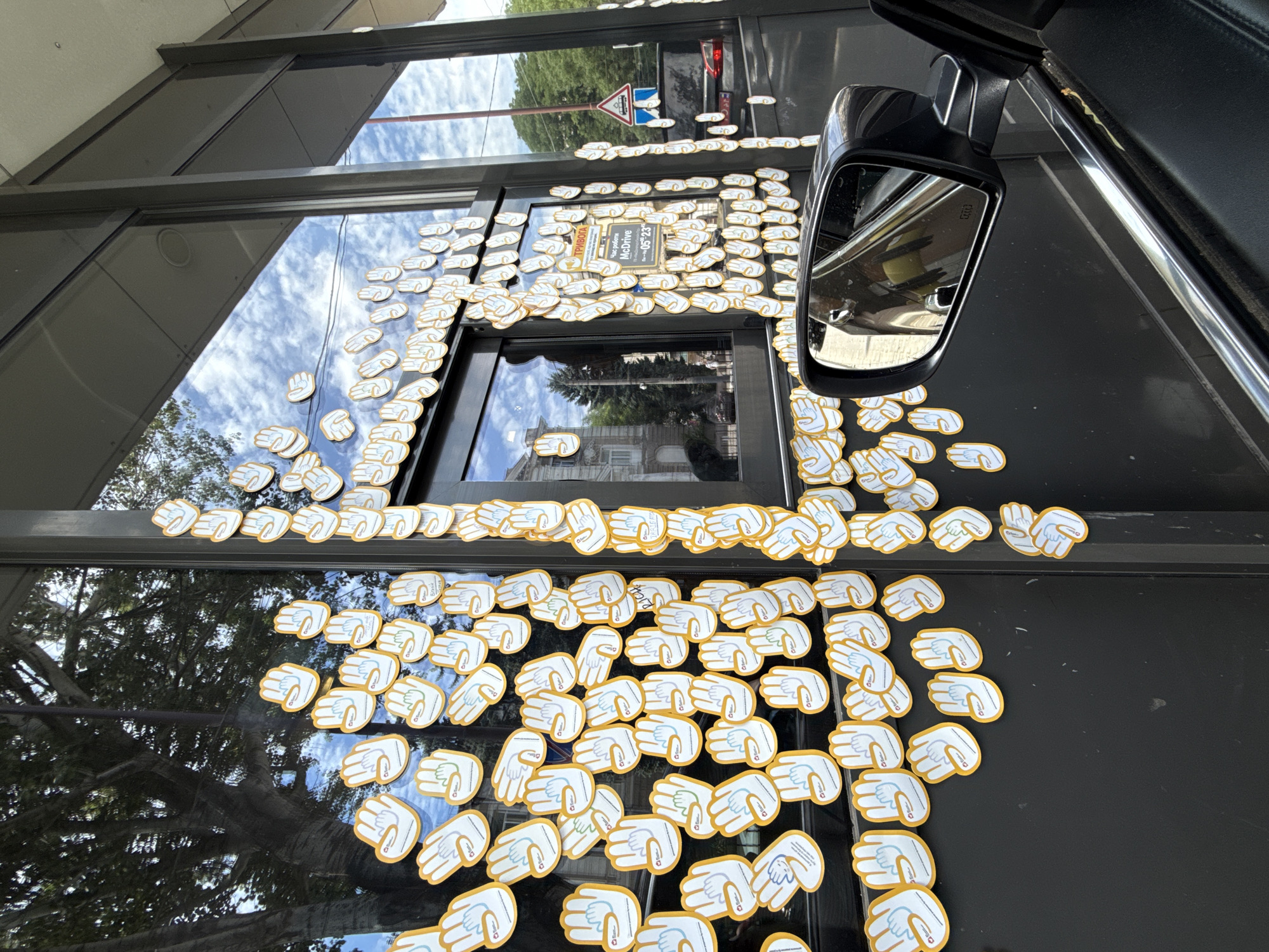 This is a photograph of a McDonald's drive-thru window decorated with numerous hand-shaped stickers. The stickers, yellow with white outlines and blue lines depicting fingers and palms, are applied densely to the exterior of the building. Some of the stickers have text in Ukrainian, including "Тривога" (Alarm) and "Час роботи МакДрайв 05-23" (McDrive Opening Hours 05-23). The window itself reflects the sky, clouds, and trees. A side-view mirror of a car is visible in the foreground, reflecting another car. In the background, trees, buildings, and a traffic sign with a truck symbol are visible. The overall setting appears to be a public area in Odesa, Ukraine, likely on a sunny day. The image captures a moment of interaction between the car and the drive-thru service.