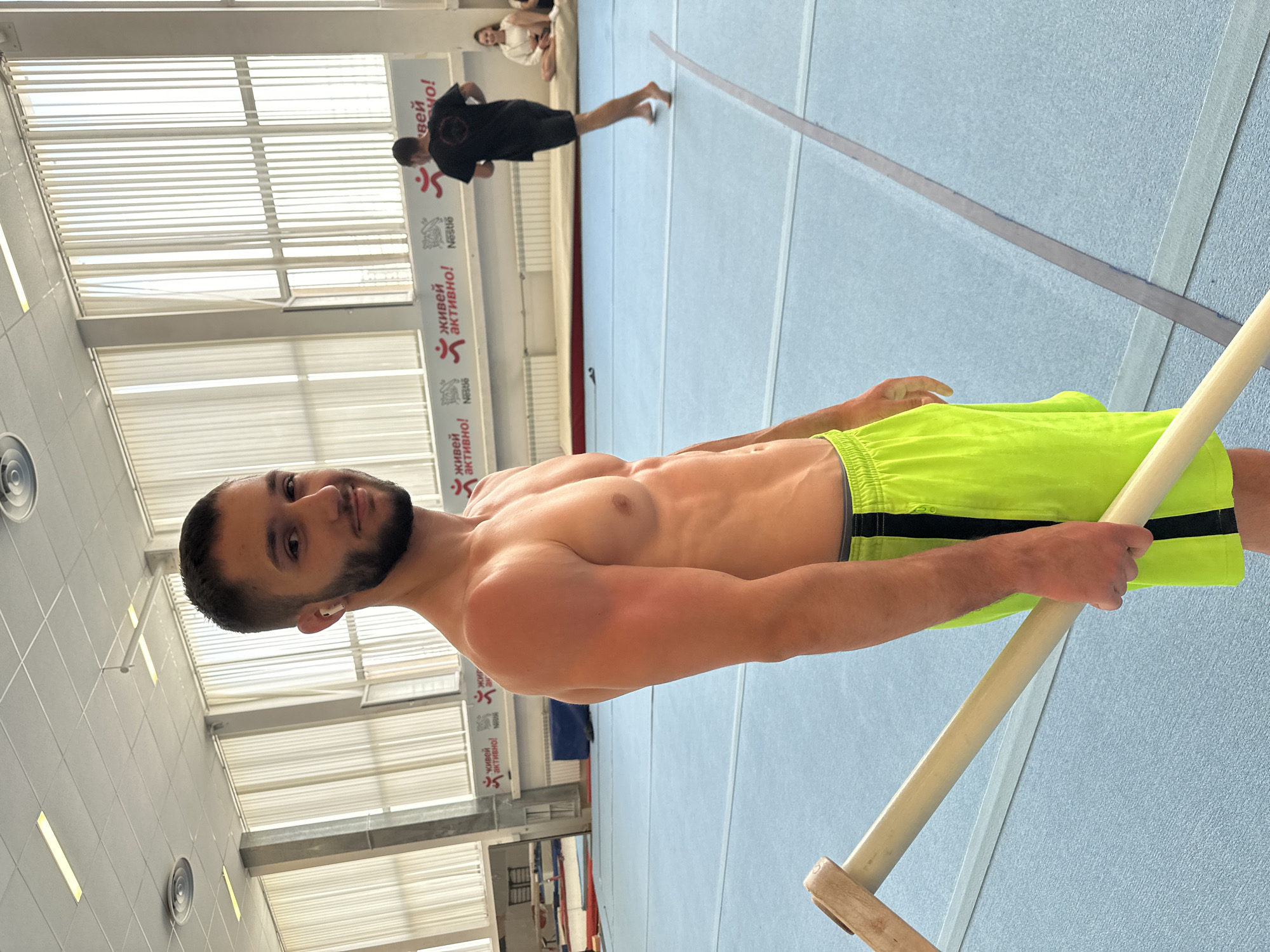 A medium eye-level shot reveals a fit man with a beard, posing shirtless in a gymnasium. He is wearing bright yellow shorts with black trim and holding a wooden gymnastics bar with both hands.
In the background, another man is captured mid-air, likely practicing gymnastics, and further back, another person is seated. The walls of the gymnasium are adorned with banners featuring text in Cyrillic alphabet and the Nestle logo. There's abundant natural light streaming in through the windows. The image has been rotated 90 degrees clockwise.
There is no evidence in the image to suggest the specific location in the city of Sofia, Bulgaria.