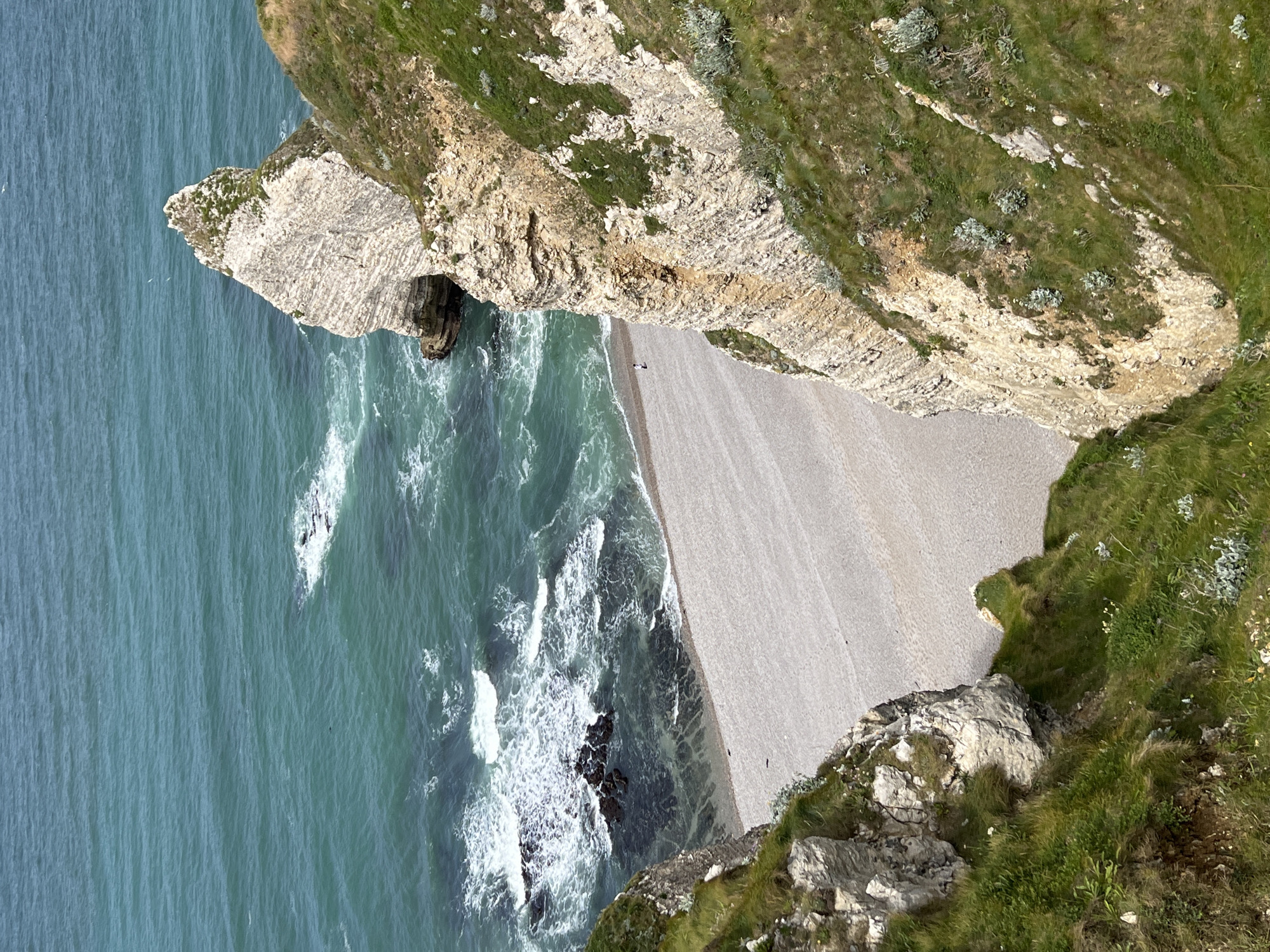 This is a high-angle, full-shot photograph taken from a cliff overlooking a beach and the ocean in Étretat, France. The sea is a vibrant turquoise with white-capped waves crashing against dark rocks and a light-colored, pebbly beach. To the right, a tall, imposing white chalk cliff with visible strata rises from the sea and slopes upwards, covered in patches of green grass and sparse vegetation. A small, solitary figure is visible on the beach, walking near the water's edge. The coastline is dramatic, with sheer cliffs and an arch-like rock formation visible in the distance on the left. The overall scene is one of natural beauty and coastal grandeur.