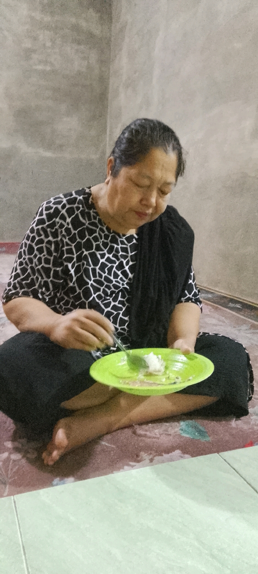 An adult woman is sitting on the floor in a room, cross-legged, eating from a green plate with a fork. She is wearing a black and white patterned shirt and black pants, with a dark shawl draped over her shoulders. The floor is covered with a patterned rug, and the wall behind her is textured and gray. A portion of a tiled floor is visible in the foreground. The woman has her eyes closed and appears to be focused on her meal. The setting suggests a domestic indoor environment, consistent with a residential area in Lenggadai Hilir, Indonesia.