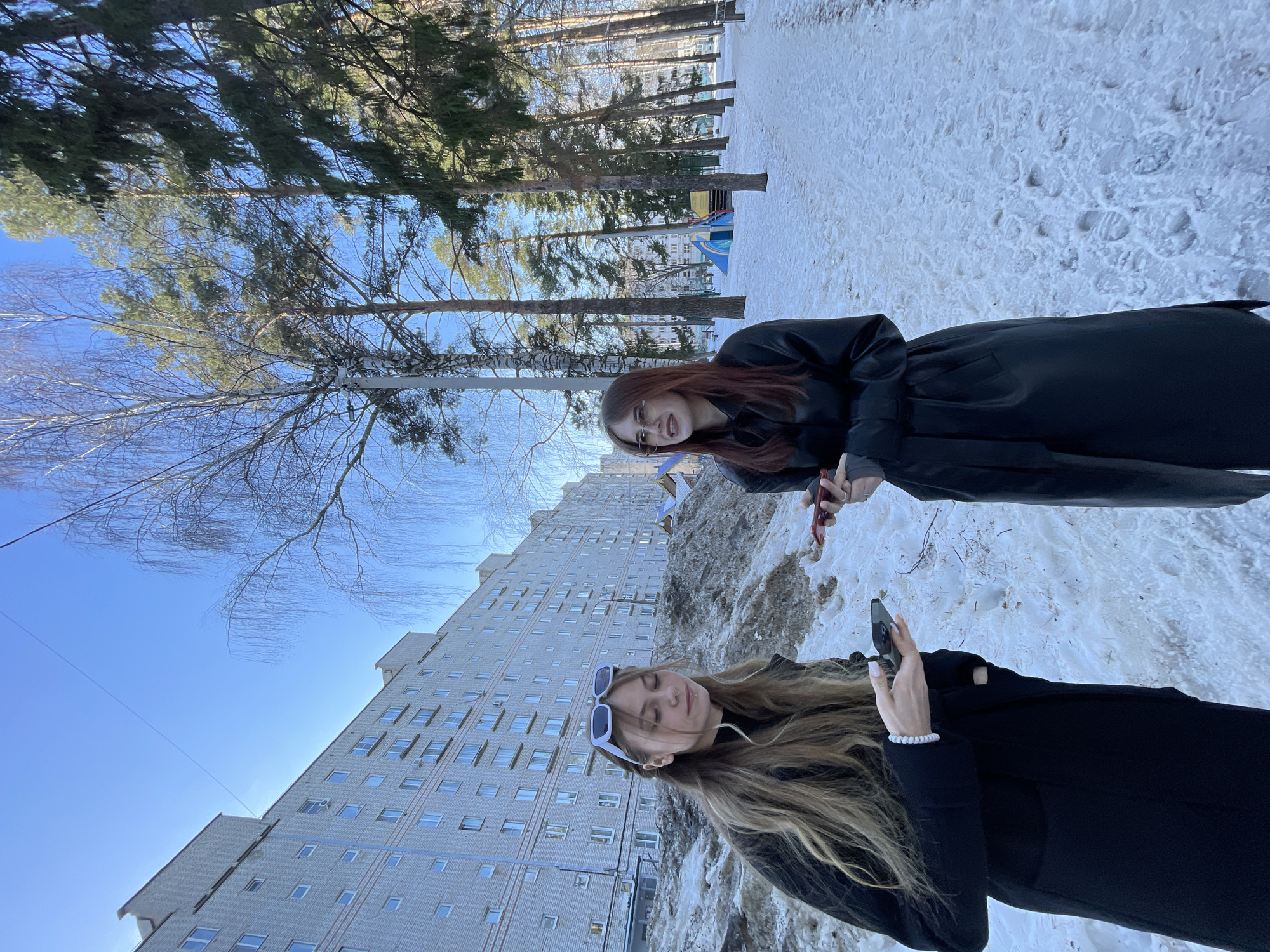 The scene depicts two young adult females in a snow-covered outdoor residential area of Chaikovsky, Russia, under a clear blue sky.
The female on the left has long, light brown hair, wears a dark coat, and has white-framed sunglasses resting on her head. She is holding a dark-colored smartphone in both hands and appears to be looking down at it. A white beaded bracelet is visible on her right wrist.
The female on the right has long, reddish-brown hair and wears clear-framed eyeglasses. She is dressed in a dark, possibly leather, long coat and holds a red-cased smartphone in her right hand. She is looking towards her left.
The ground is covered with snow, interspersed with patches of exposed dirt and ice, particularly along a path visible on the right. In the background to the left, a large, multi-story apartment building constructed of light-colored brick is prominent. To the right and center-background, numerous tall evergreen trees and bare deciduous trees are visible. A children's playground structure with blue and yellow components can be seen among the trees in the mid-ground. A thin wire spans the upper portion of the sky.