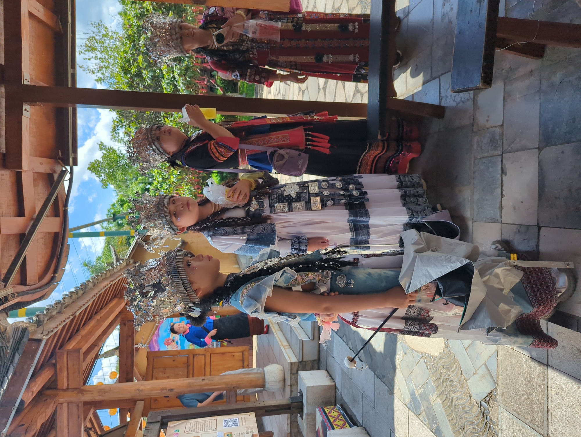 In Xishan District, China, four individuals are standing under a wooden canopy, likely a part of an outdoor market or cultural attraction. The subjects are dressed in traditional ethnic attire, characterized by ornate headpieces, intricate patterns on their clothing, and layered fabrics. The individuals appear to be women or girls, with varying ages suggested by their features.
The person on the far left, a young girl, is facing to the right and holding a white object, possibly an umbrella or a prop, with a stick. Her attire is a mix of blue and white, with decorative silver elements and beaded braids. To her right, another young girl is also looking to the right, holding a yellow cup and wearing a white and blue dress with similar traditional ornamentation.
The third person from the left is a woman wearing a black and colorful patterned outfit. She is holding a yellow cup and has a purple bag slung over her shoulder. The woman on the far right is dressed in a striking red and patterned garment. She is holding a plastic bag with some items inside and appears to be looking towards the left.
The environment suggests a sunny day with a partly cloudy sky visible in the background. Lush green trees are present, indicating a natural setting or well-maintained landscaping. The ground is paved with stone tiles. Wooden structures, including beams and a roof, are visible, suggesting a sheltered area for vendors or visitors. The overall scene evokes a cultural and traditional atmosphere.