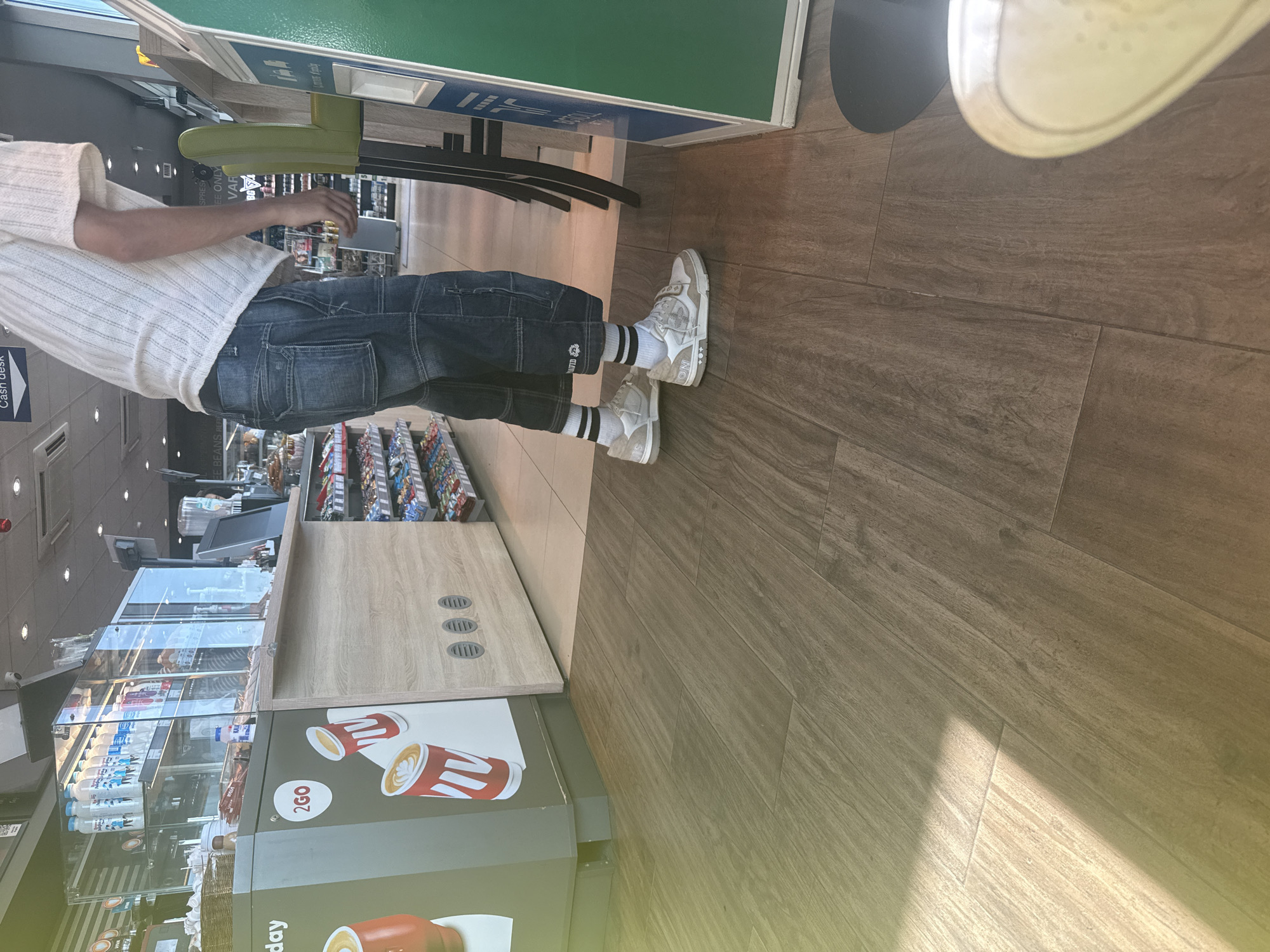 The image depicts the interior of a retail establishment, likely a convenience store or gas station, with a focus on a person standing near a counter. The flooring is a wood-effect tile. The person is wearing a white, textured short-sleeved shirt, dark blue denim capri pants with multiple pockets, black and white striped socks, and white athletic shoes. The counter area features a display of bottled drinks in a refrigerated case, a coffee machine, and promotional materials for "2GO" coffee. Shelves stocked with packaged goods are visible in the background. A green upright sign is positioned to the right of the person. The scene is brightly lit. The location is identified as Drazhevo, Bulgaria.