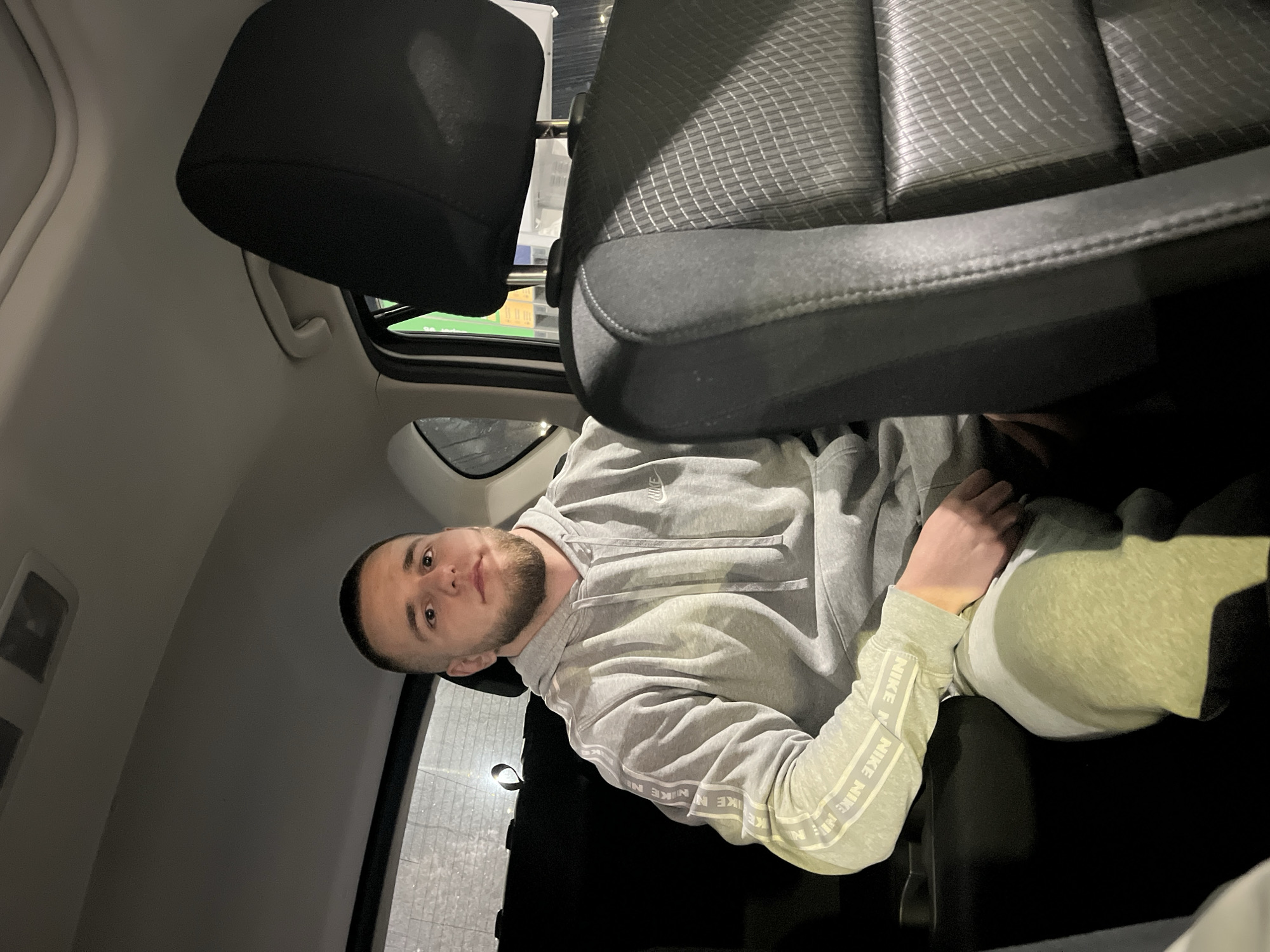 A person with a beard is sitting in a car, wearing a gray hoodie with "NIKE" repeating down the sleeves. The view is from the back seat looking towards the front passenger seat. The interior is dark, but there's a sunroof with a glimpse of an illuminated exterior that could indicate an urban environment. There is no information to identify a specific location in Burgas, Bulgaria.