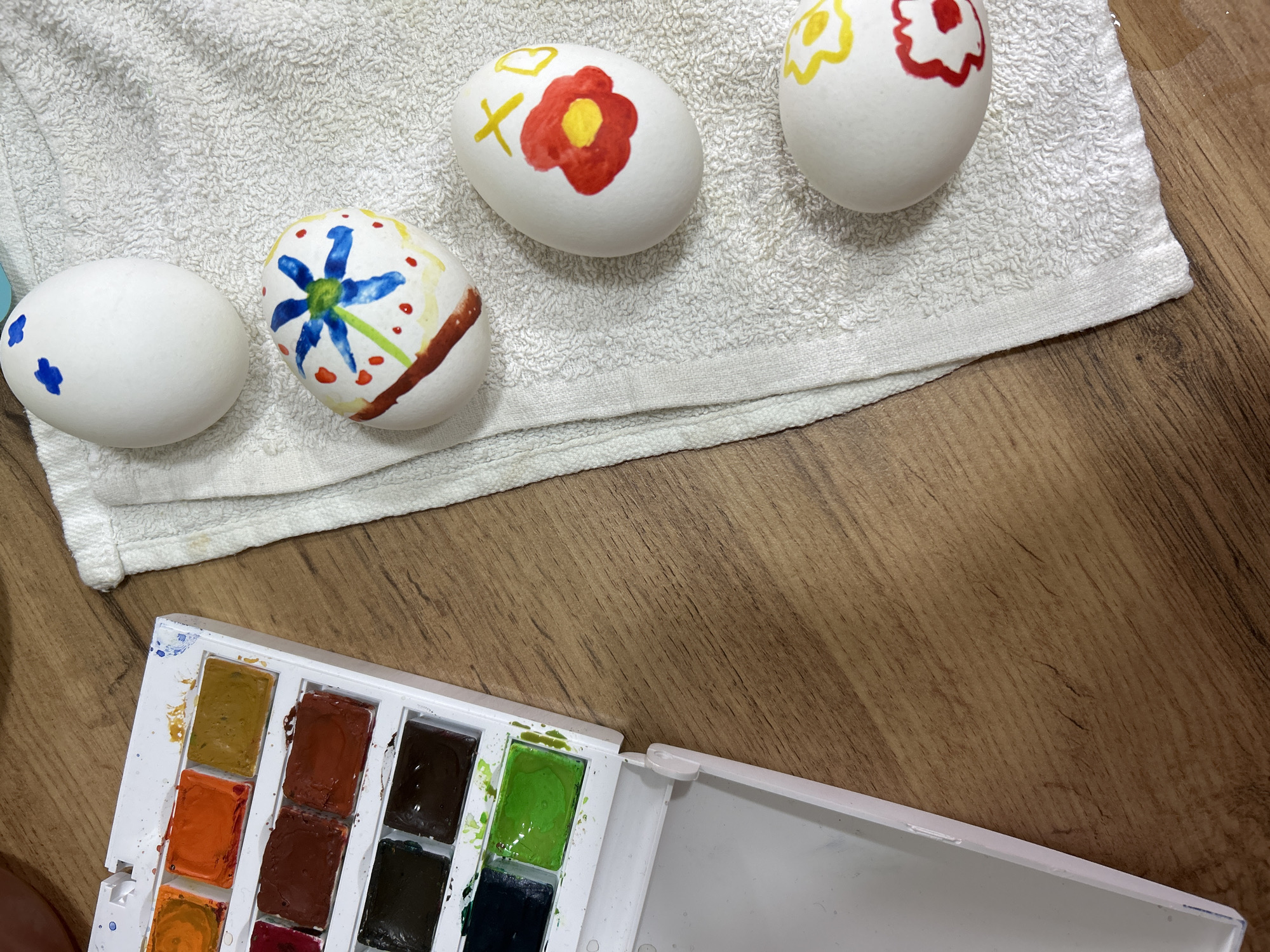 The image shows four painted Easter eggs on a white towel, with a watercolor paint set in the foreground. The eggs are decorated with different designs using watercolor paints, including a flower, a cross, and a few dots. The towel is on a wood-grain surface, presumably a table. The watercolor paint set appears to be in a plastic container and contains various colors.
The location is unknown. The city and country were not provided in the image.