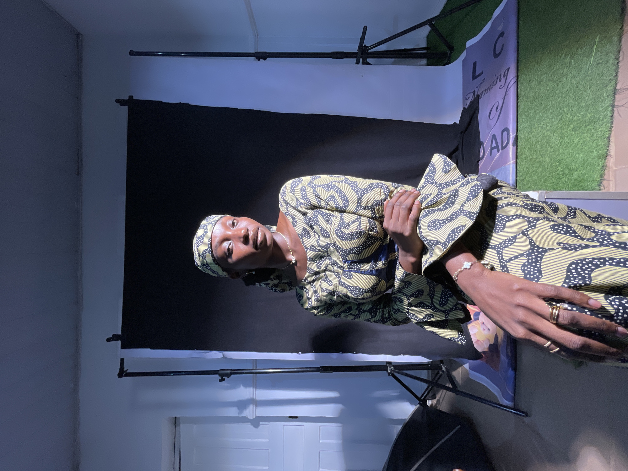 This image captures a posed portrait of a Black woman within an indoor photography studio setting in Jos, Nigeria. The woman is the central subject, standing and looking directly at the camera with a calm, neutral expression. She is adorned in a vibrant outfit, comprising a top/dress and a matching headscarf, all featuring a distinctive pattern of light yellow/beige with black squiggly lines and white polka dots. Her accessories include a delicate gold chain necklace, a gold bracelet with a white four-leaf clover charm on her right wrist, and multiple gold rings on her fingers, which are clasped together in front of her.
The background setup suggests a temporary or dedicated studio space. Behind the woman is a large black backdrop, supported by visible stands. To her immediate right, a banner with partially visible text is displayed against a wall covered in artificial green grass. Visible text on the banner includes "LC", "Fostering Of", and "AD", potentially followed by more letters. The banner's background is a deep blue or purple with white lettering. To her left and behind her, a plain white wall is visible. The scene is illuminated by strong artificial lighting, creating prominent highlights on the woman's face and clothes, and casting distinct shadows on the white wall, emphasizing the dramatic studio environment. The overall activity is a formal portrait photography session.